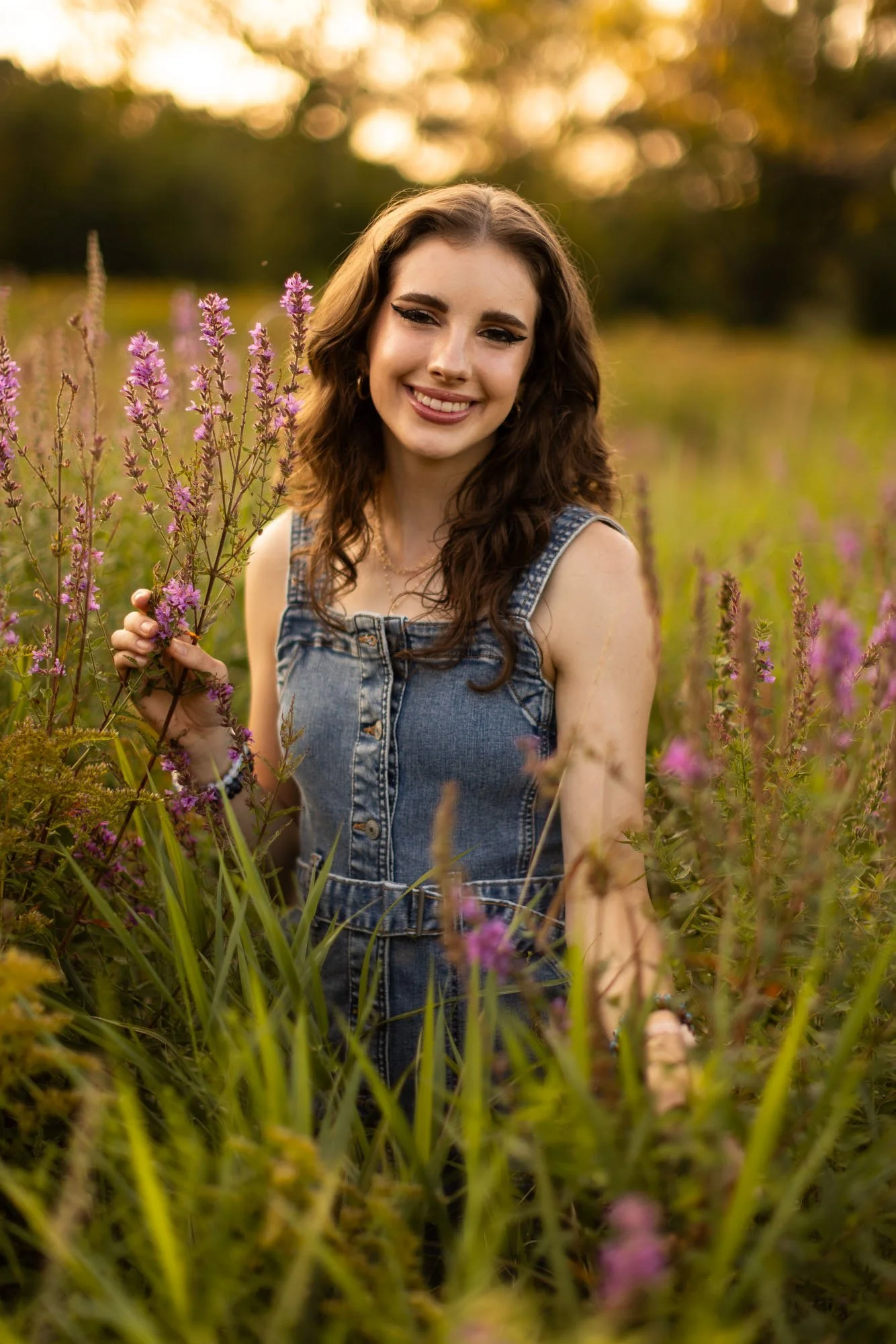 Smiling young woman with wavy brown hair holding purple flowers in a field of tall grass and purple flowers during sunset.