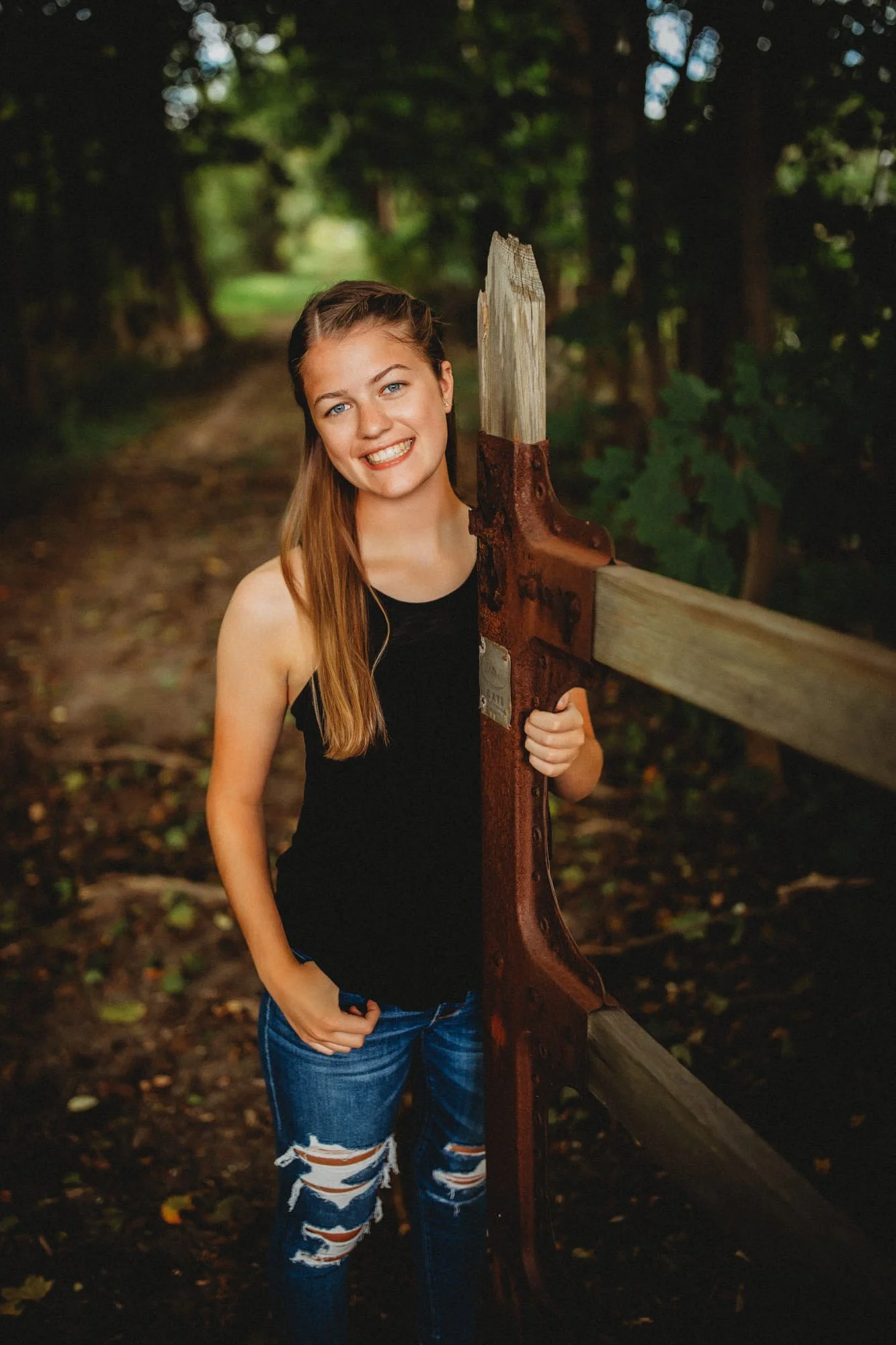 A young woman with long, light brown hair, smiling and holding a large weathered metal and wood crowbar, standing on a forest trail with green trees and foliage in the background.