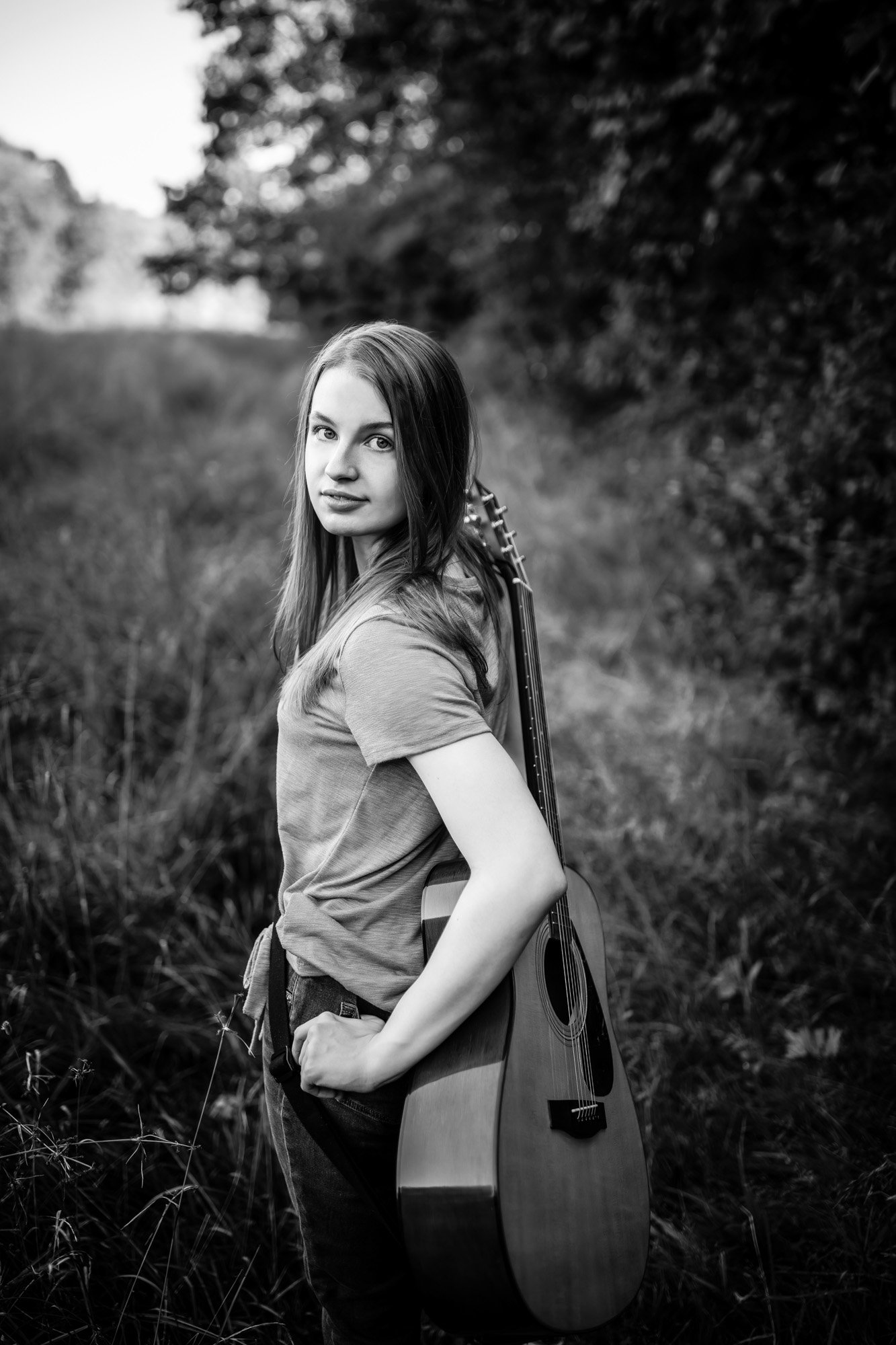 A young woman standing outdoors in a natural setting with tall grass and trees in the background, holding an acoustic guitar over her shoulder, looking at the camera in black and white.