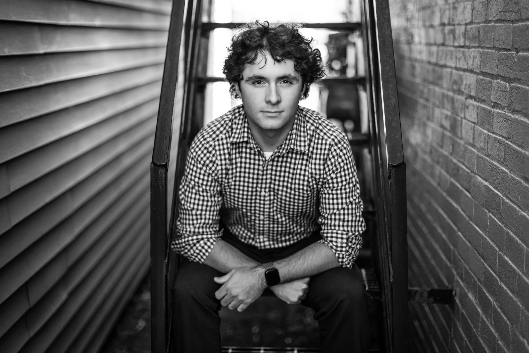 A young man with curly hair and a checkered shirt sitting on stairs between a brick wall and siding, looking at the camera.