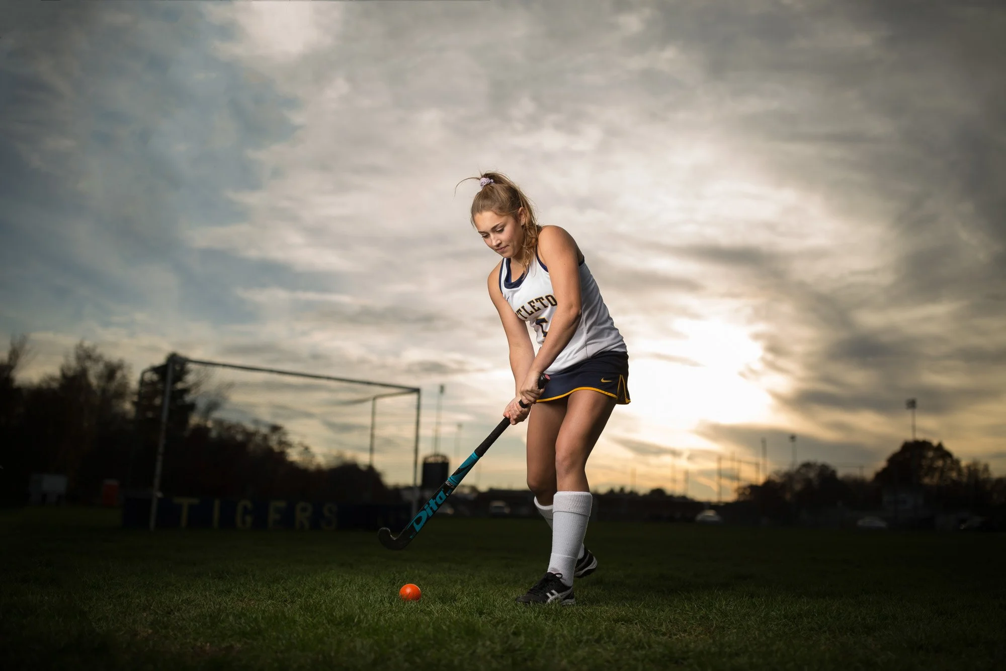 Female field hockey player in uniform preparing to hit an orange ball on a grassy field during sunset.
