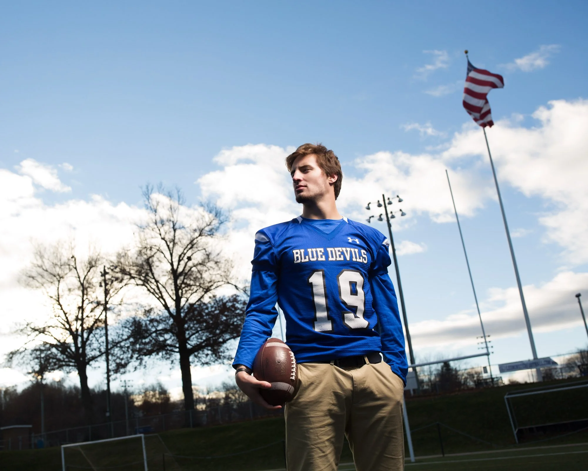 A young man in a blue football jersey with number 19 and 'Blue Devils' printed on it, holding a football, standing outdoors on a sports field with flags, trees, and a partly cloudy sky in the background.