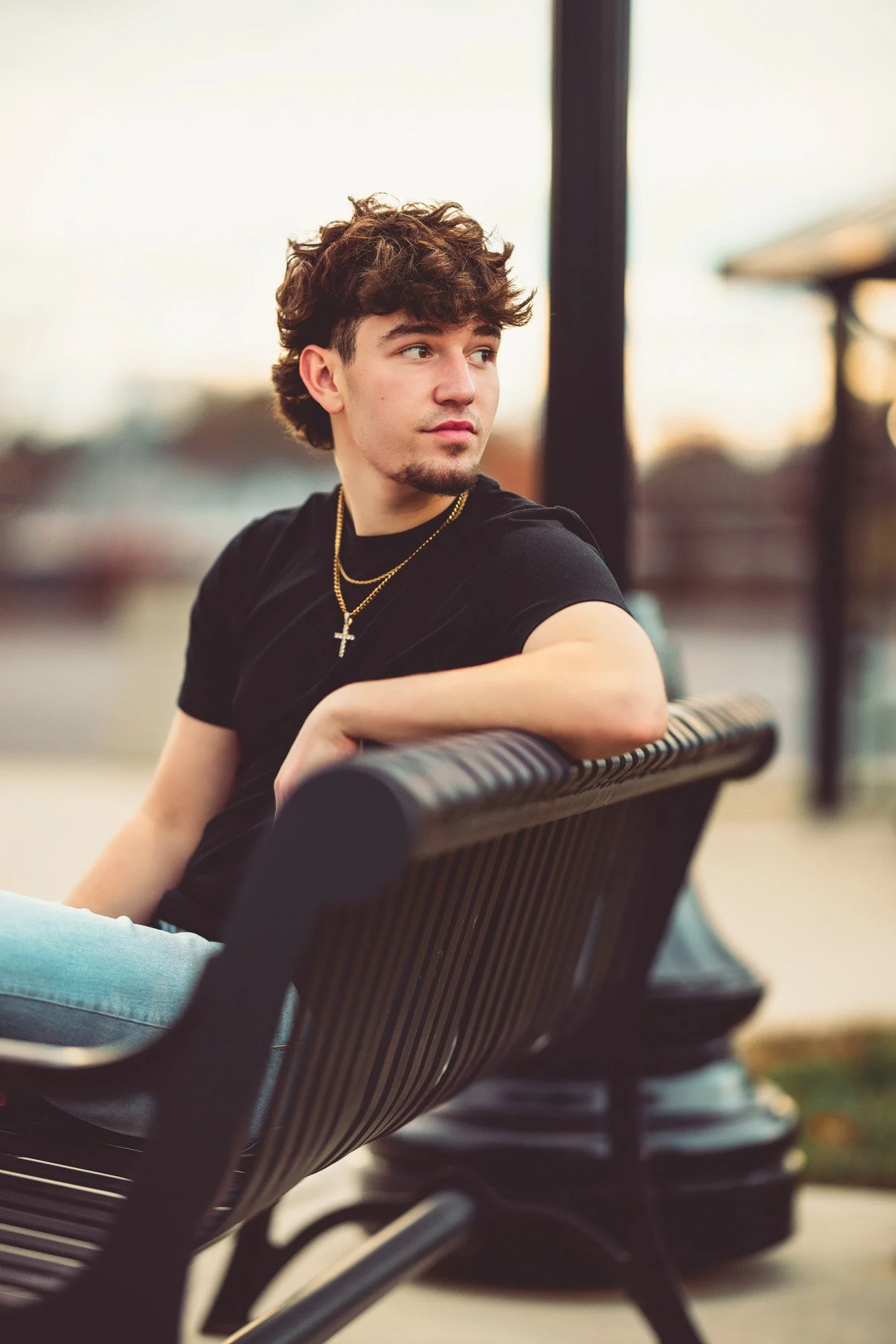 Young man with curly brown hair sitting on a park bench, wearing a black t-shirt and gold chains, gazing into the distance during sunset.