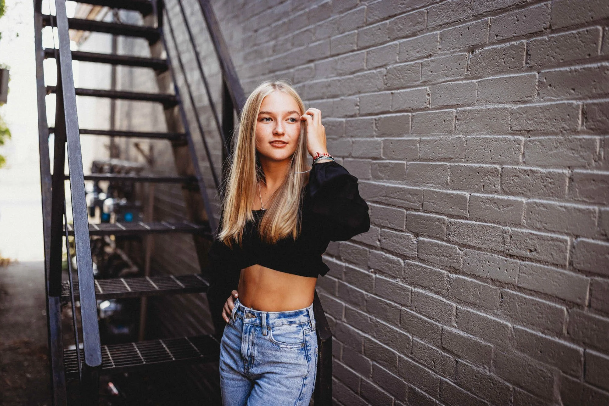 A young woman with long blonde hair leaning against a brick wall, touching her face with one hand, standing next to an outdoor metal staircase.