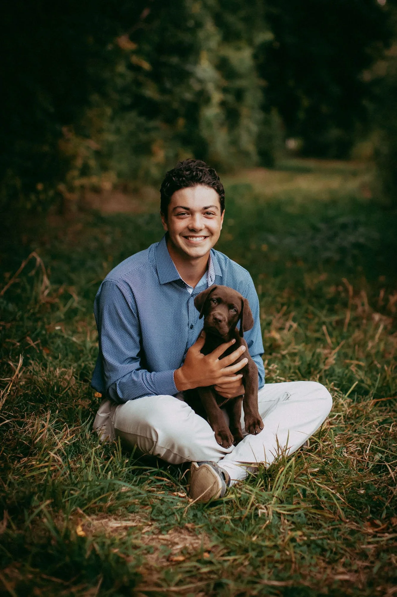 A young man sitting on the grass outdoors, holding a chocolate Labrador puppy, smiling at the camera with trees in the background.