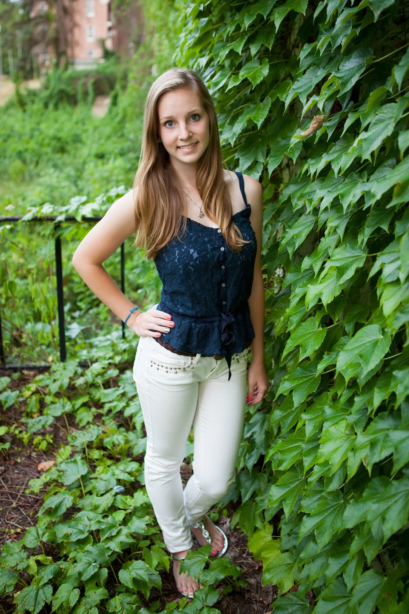 A young woman with long light brown hair, wearing a navy blue lace top and white pants, standing outdoors next to a large green ivy-covered wall and a garden with lush green foliage.