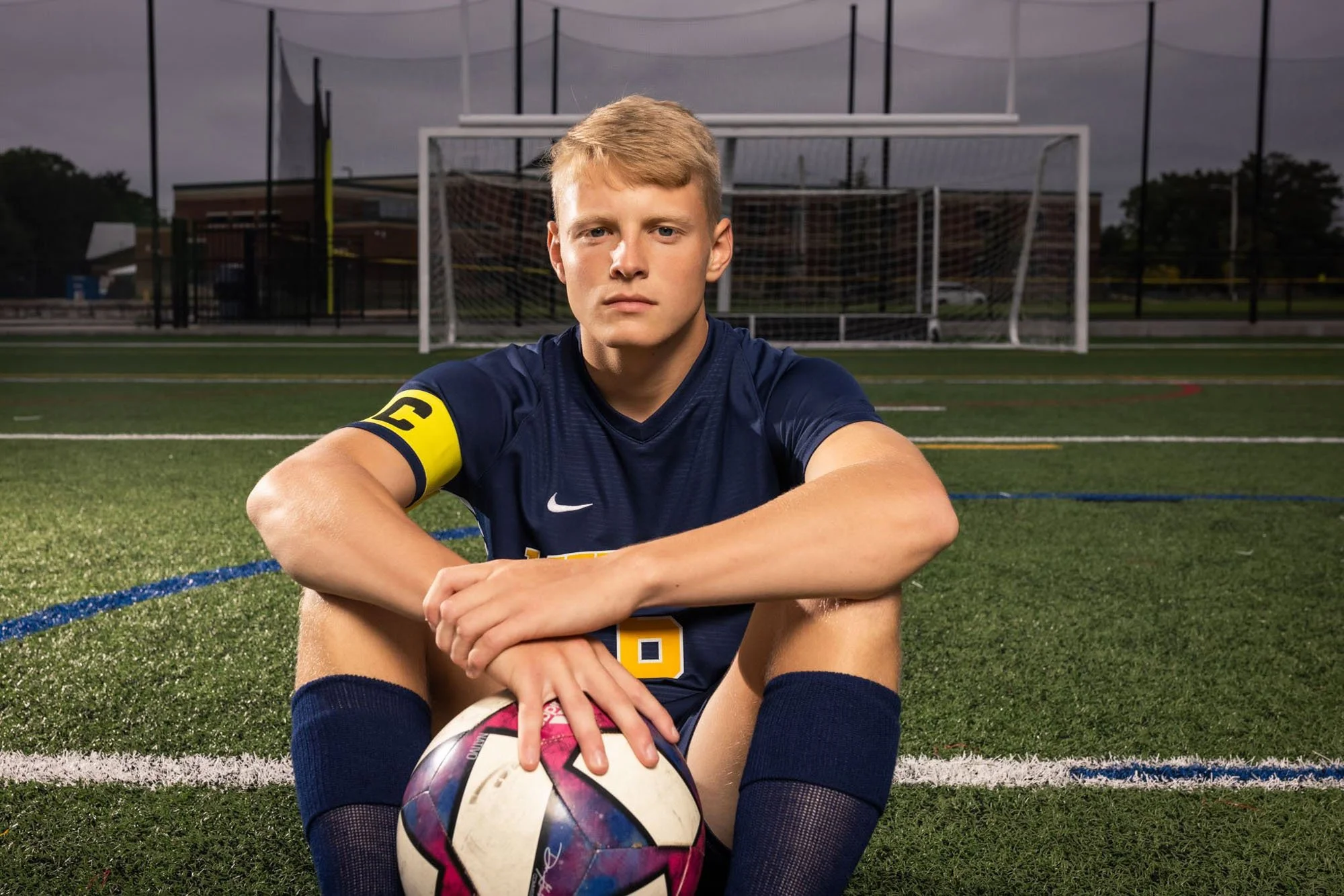 Young male soccer player sitting on the field holding a soccer ball, wearing a navy blue uniform with a yellow captain's armband, on a soccer field with goalpost in the background.