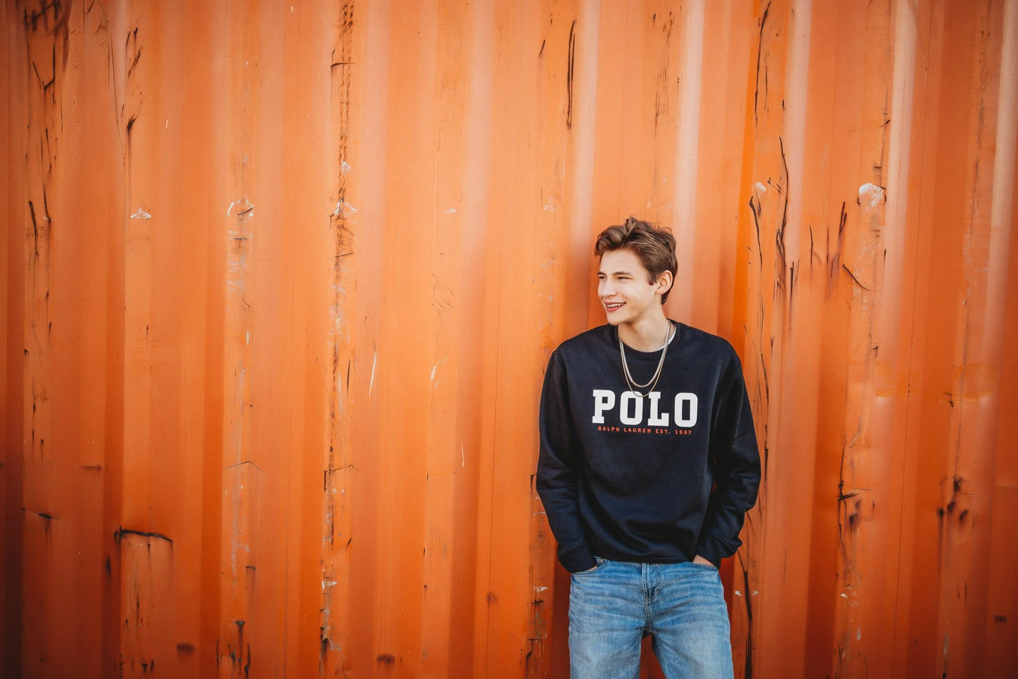 Young man smiling and standing against an orange metal wall, wearing a black sweatshirt with 'POLO' written on it and jeans.