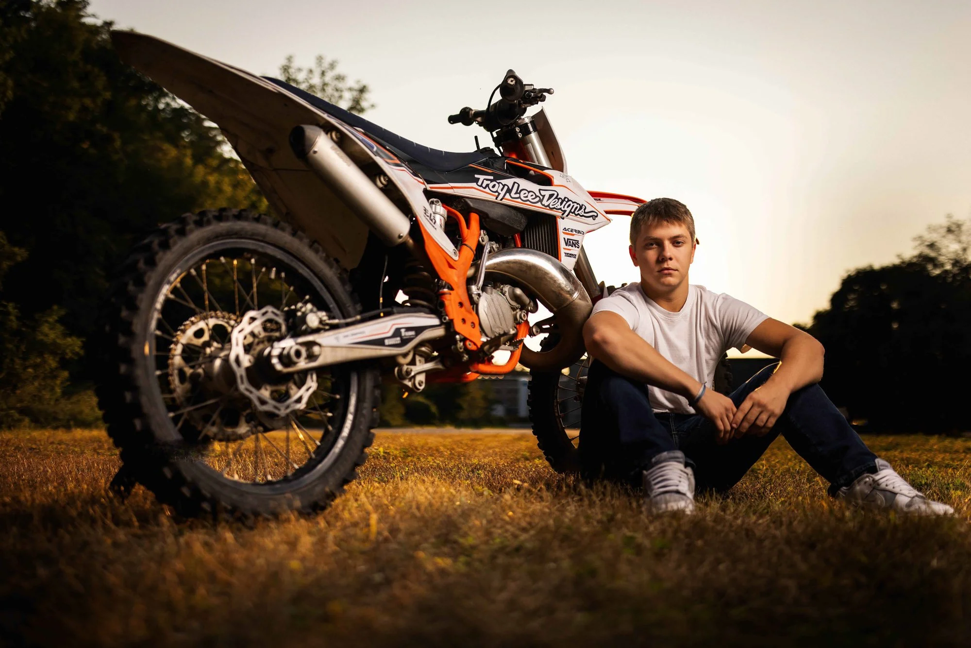 A young man sitting on the grass next to an orange and black dirt bike outdoors during sunset.