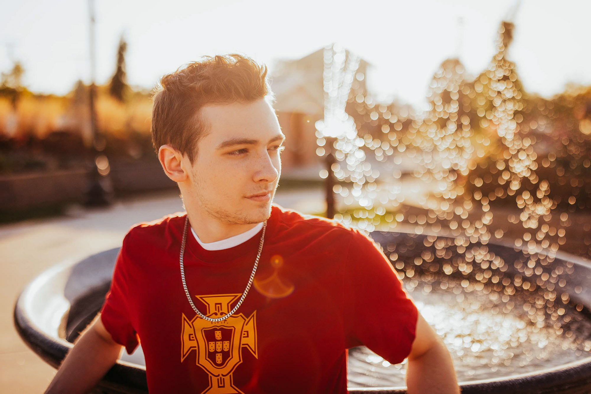 Young man with short brown hair wearing a red T-shirt with a yellow and black emblem and a silver chain necklace, sitting outdoors in the sunlight, with trees and a house blurred in the background.