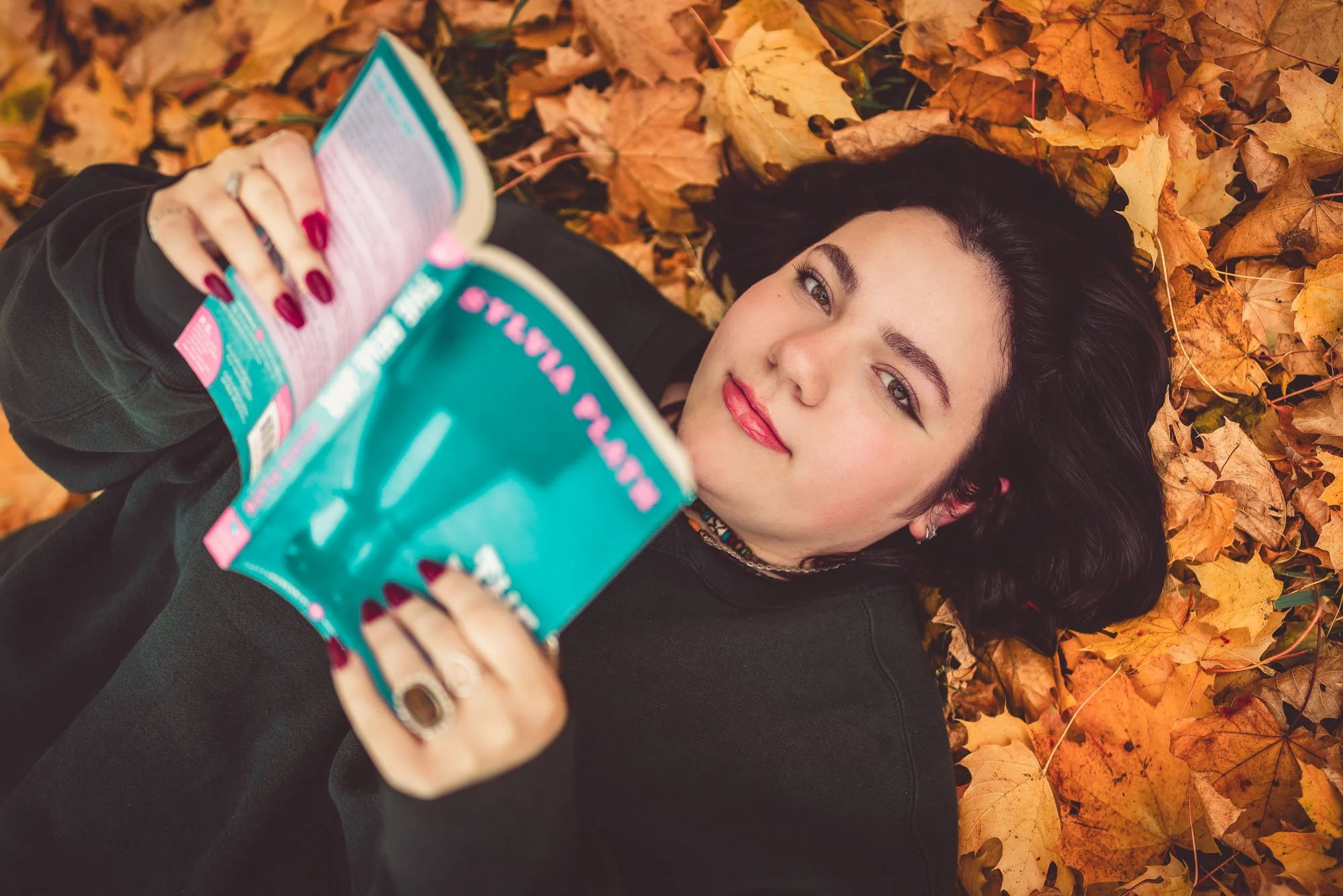 A woman with dark hair lying on a bed of orange fall leaves, holding a teal packet and looking at the camera with a slight smile.
