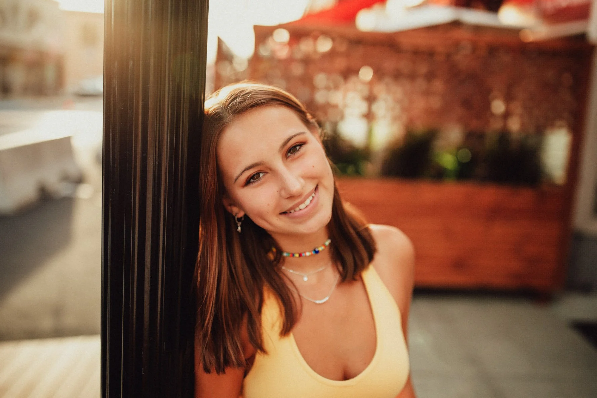 A young woman with light skin, brown hair, and a yellow top smiling and leaning against a black post outdoors during sunset.