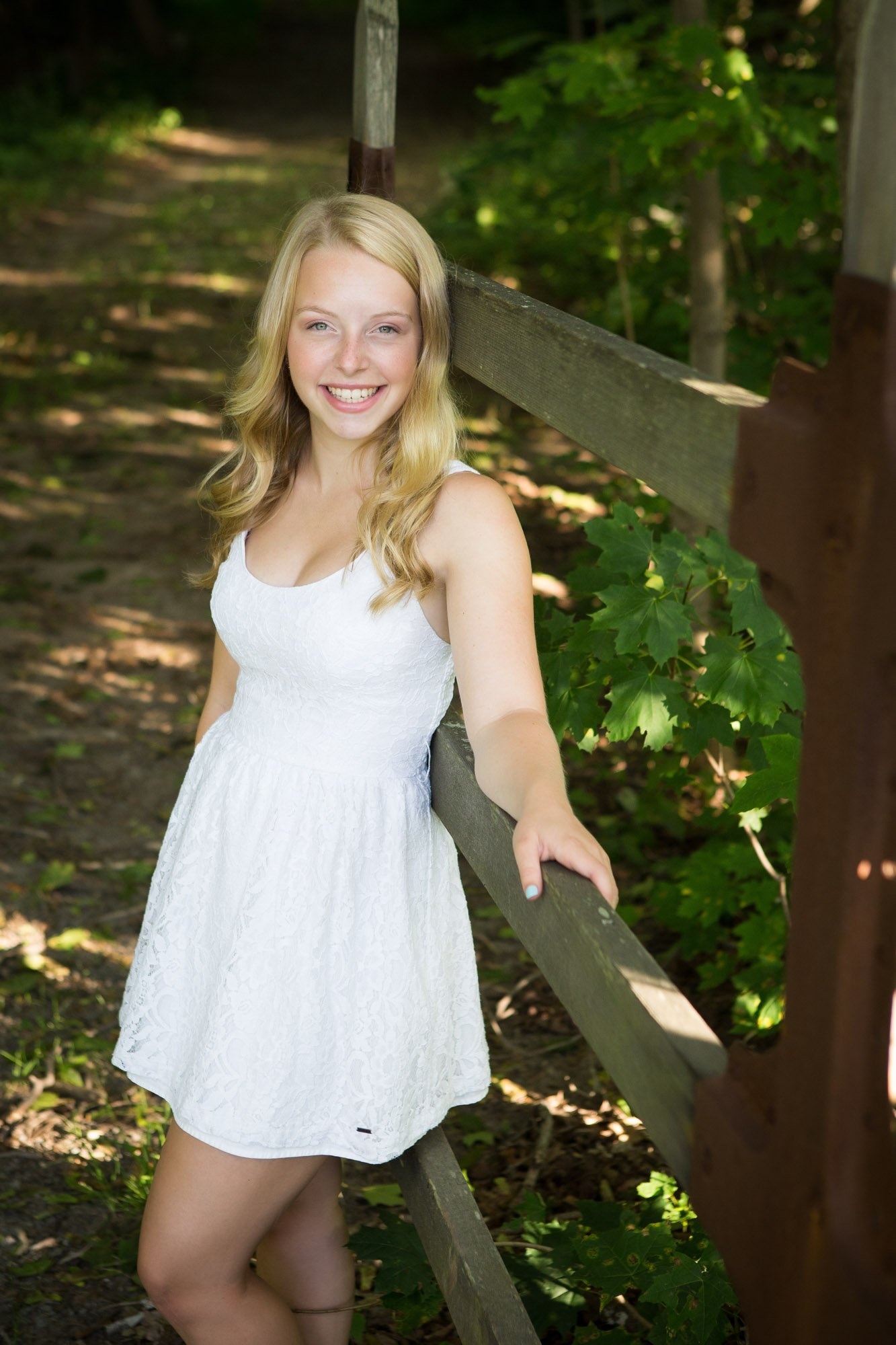 A young woman with blonde hair wearing a white dress smiling and leaning on a wooden fence in a wooded area.