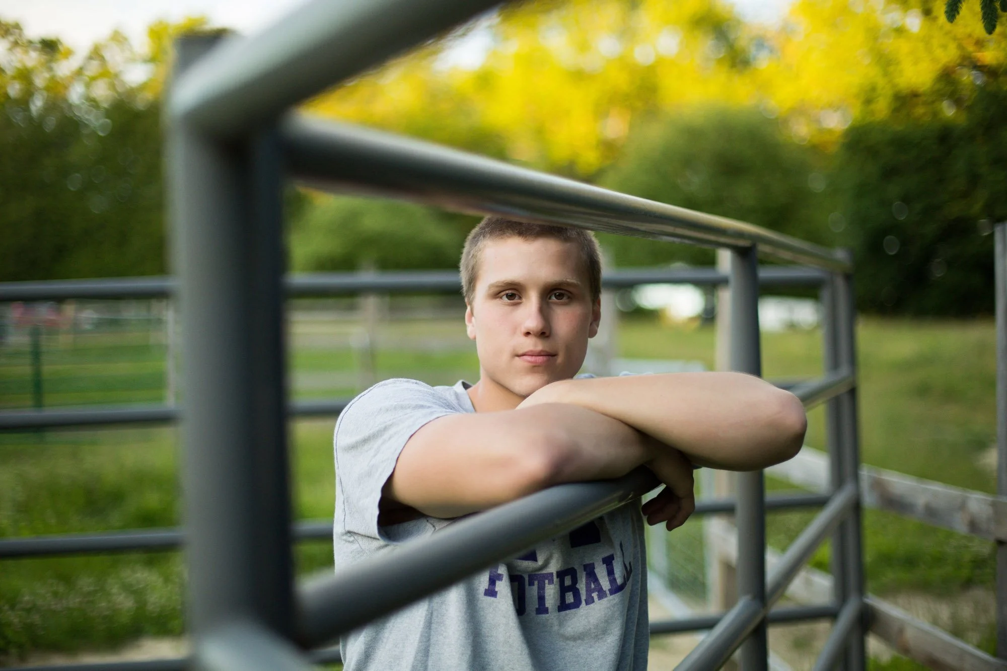 A young man with short hair leaning on a metal fence outdoors, with trees and a grassy field in the background.