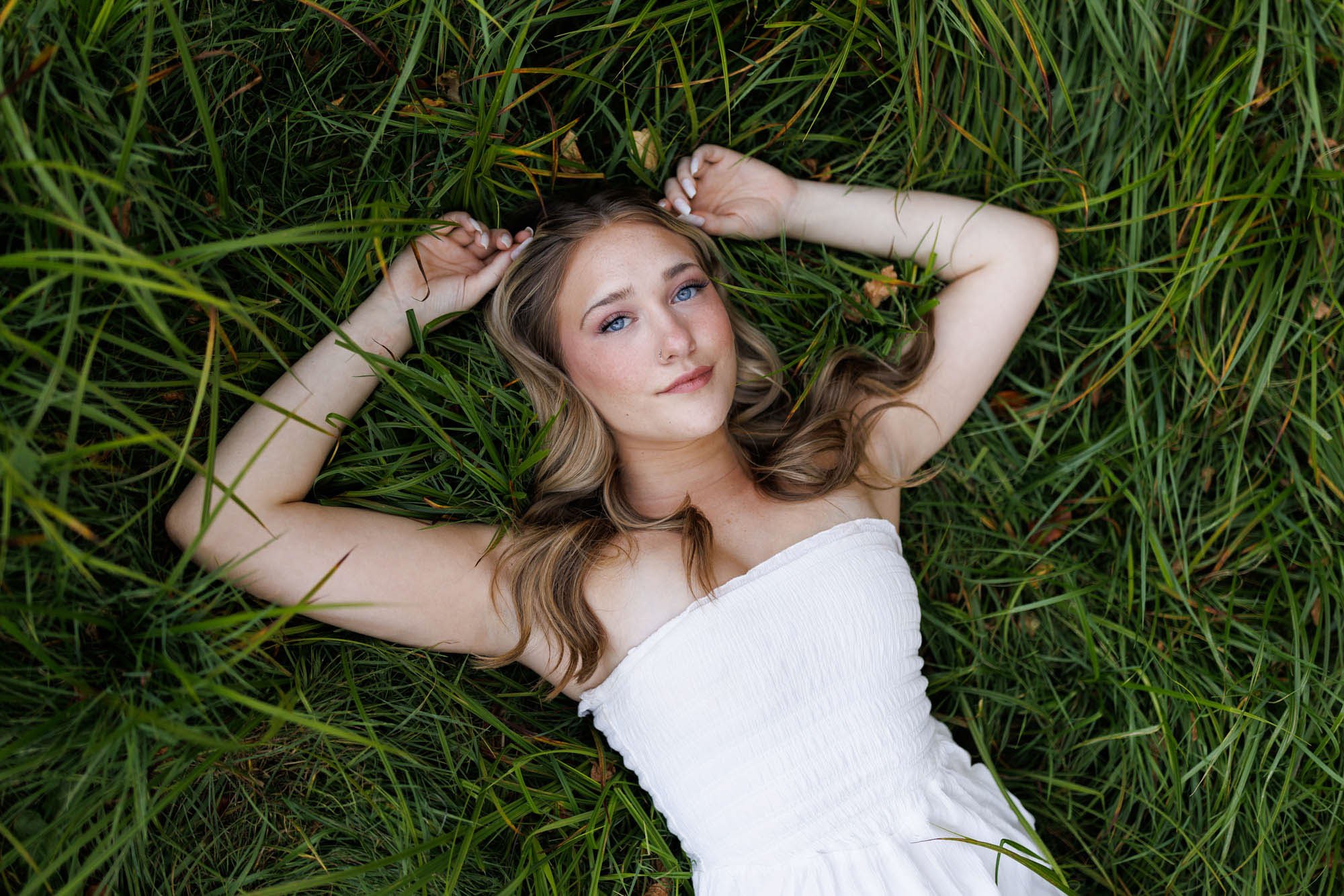 Young woman with long, wavy hair lying on green grass, wearing a white strapless dress and looking at the camera.