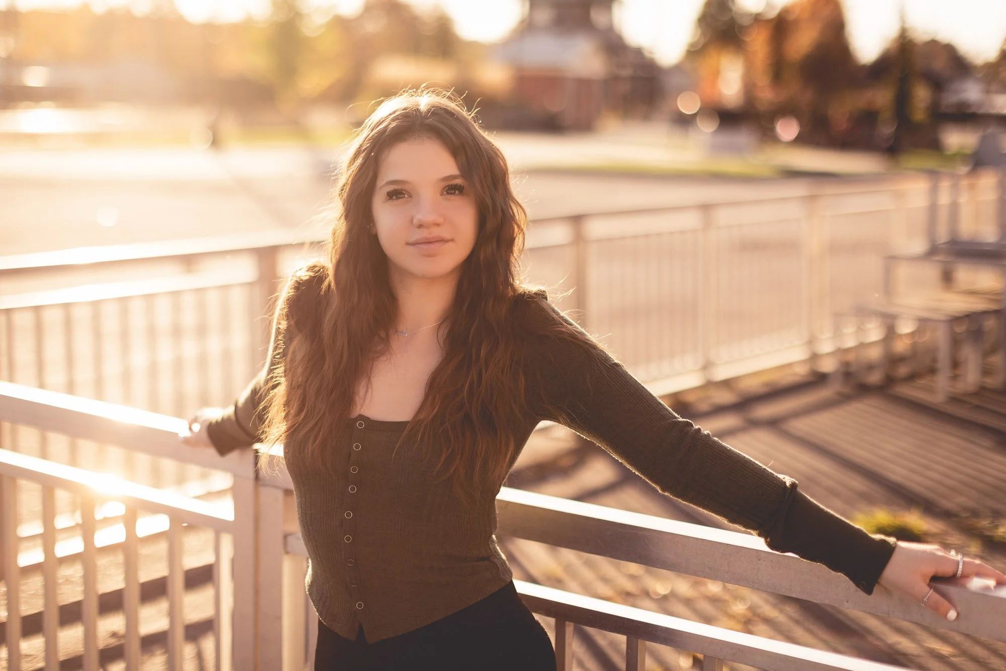 Young woman with long, wavy brown hair standing outdoors on a bridge or railing, with a blurred park or open field background bathed in warm, golden sunlight.