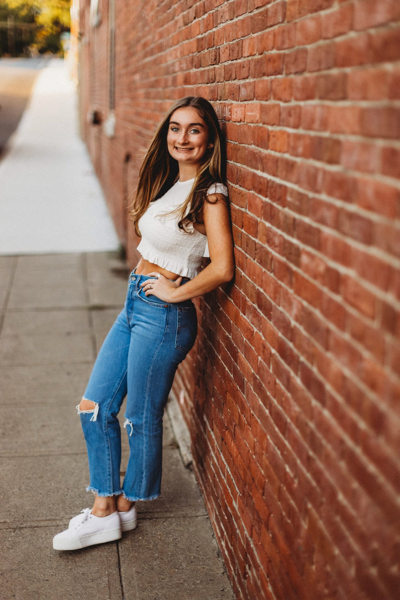 A young woman leaning against a red brick wall on a city sidewalk, smiling at the camera, dressed in a white cropped top, ripped blue jeans, and white sneakers.