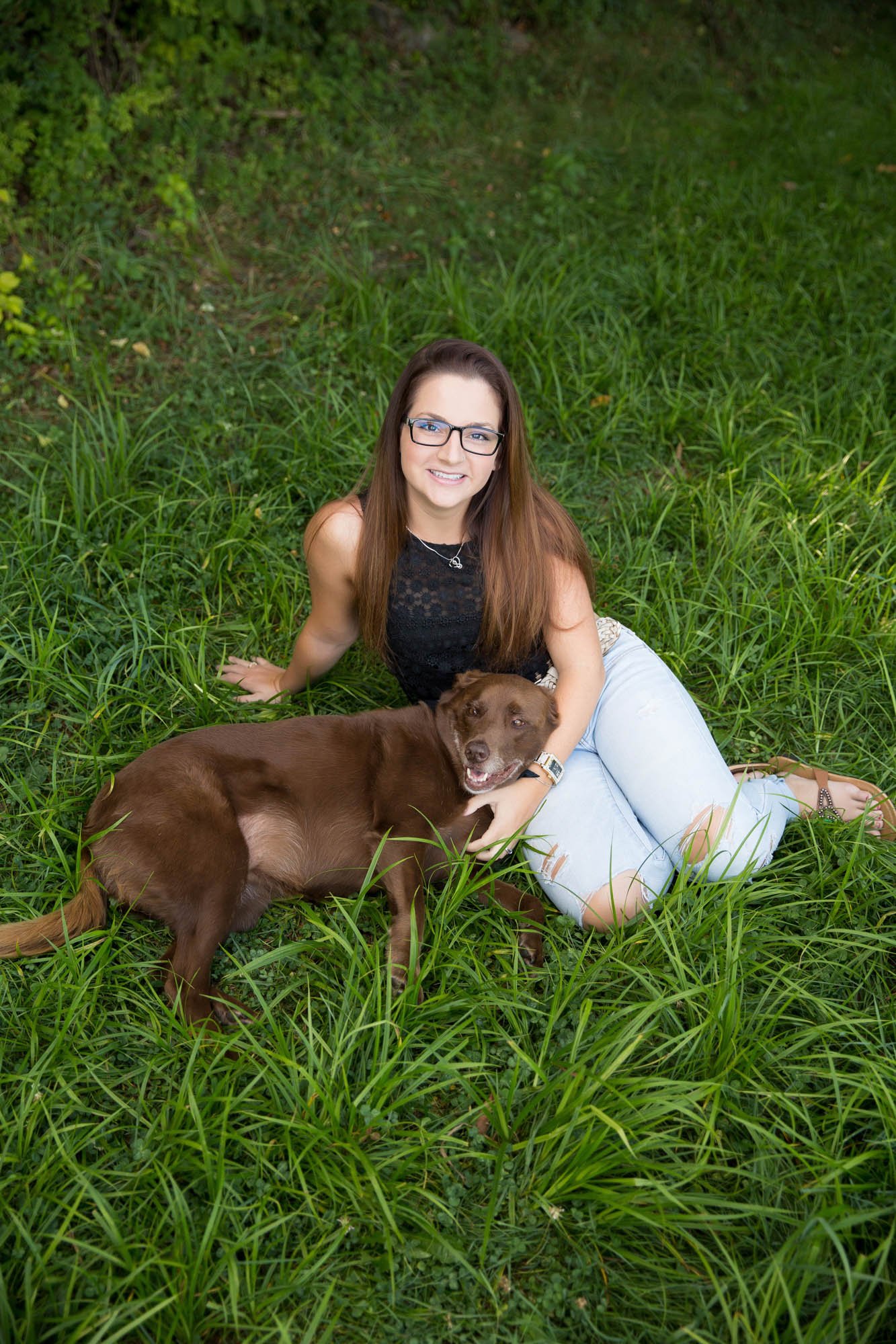 A smiling woman with long brown hair, glasses, wearing a black sleeveless top and ripped light blue jeans, sitting on green grass outdoors with a brown dog lying next to her, in a natural setting.