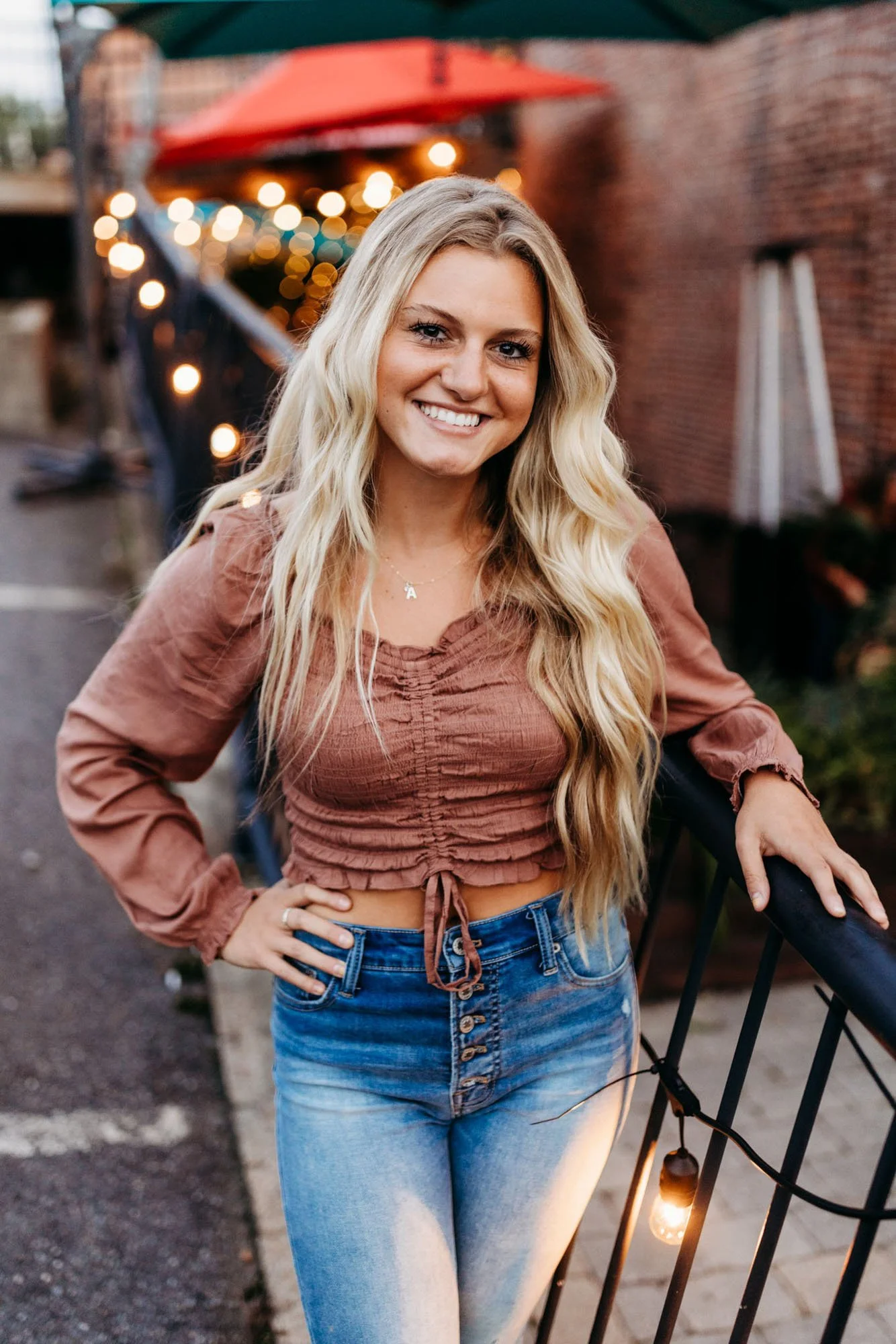 A young woman with long blonde hair smiling outdoors at dusk, standing near a black metal fence and a string of lights, with a brick wall and a red umbrella in the background.