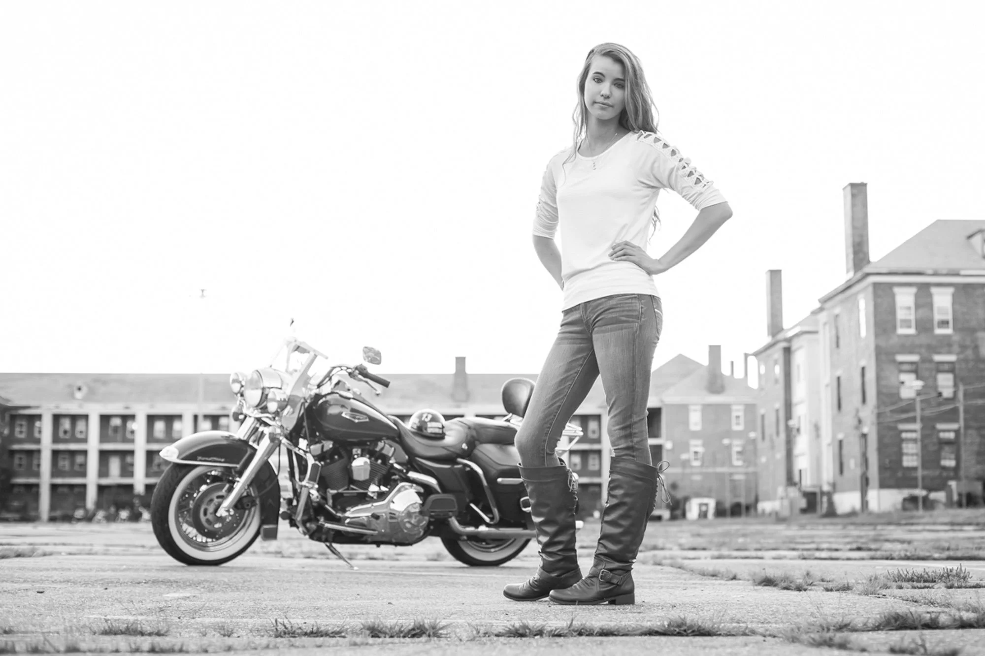 A young woman with long hair, wearing a white top with cut-out shoulders, jeans, and boots, stands outdoors with her hands on her hips. A motorcycle is parked behind her on a paved area with a background of residential buildings.