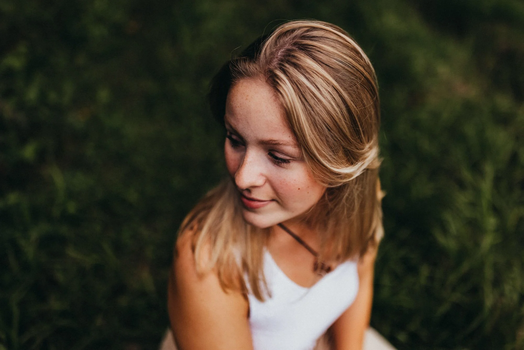 Pointed overhead shot of a smiling woman with blonde hair wearing a white tank top, sitting outdoors on green grass.