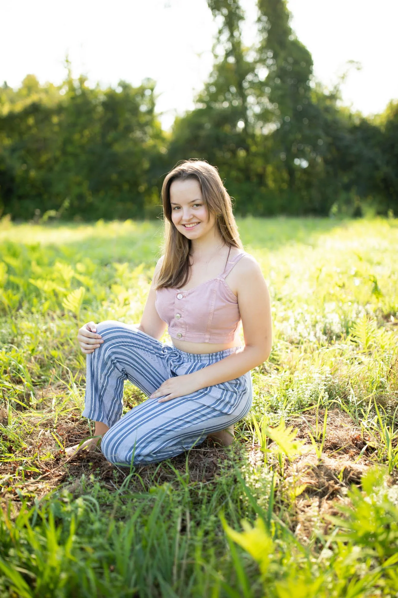A young woman with brown hair kneeling on grass in a field, smiling at the camera, with trees in the background during daylight.