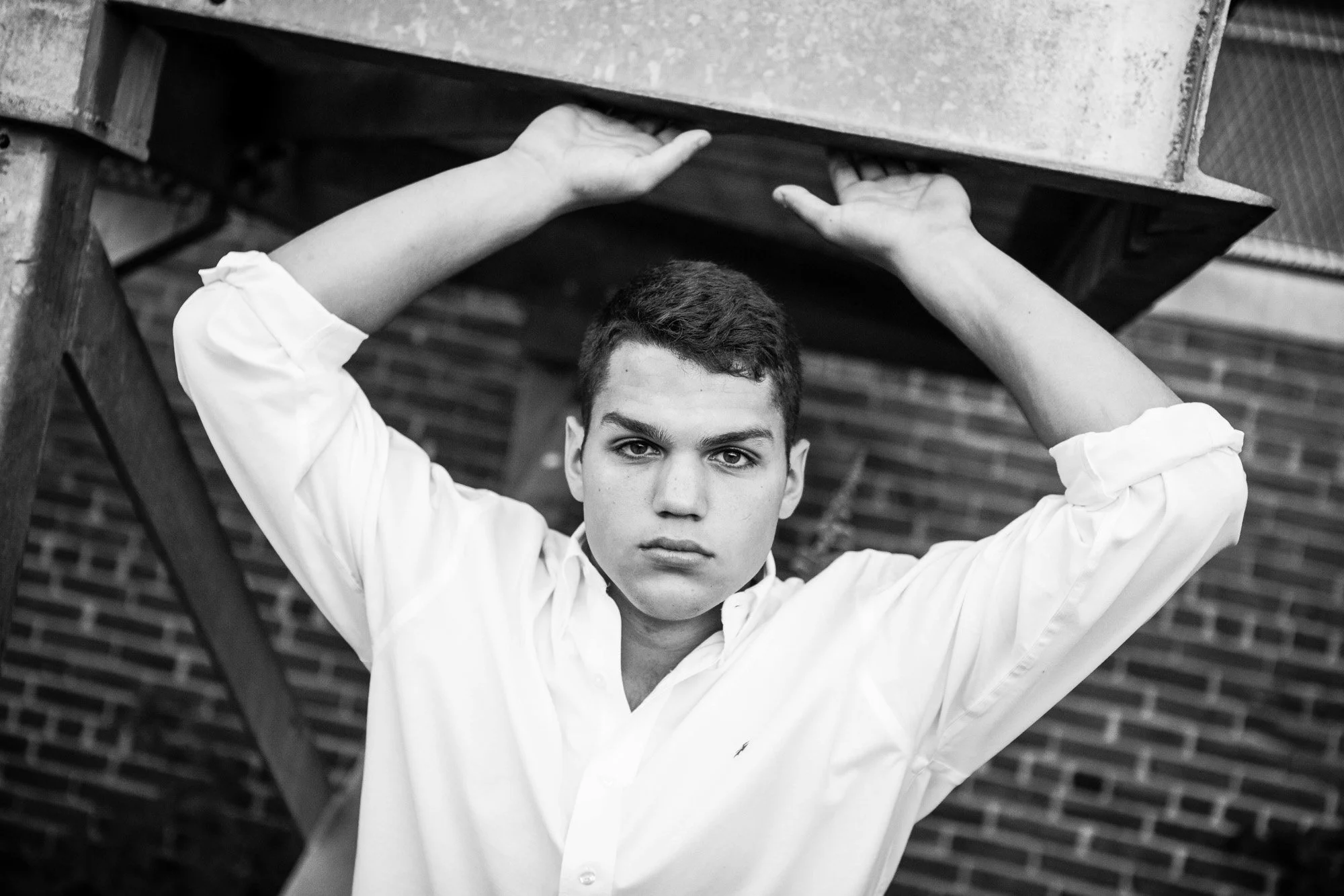 Black and white photo of a young man with short dark hair, wearing a white shirt, looking directly at the camera with a serious expression, standing outdoors in front of a brick wall, holding a metal platform above his head with both hands.