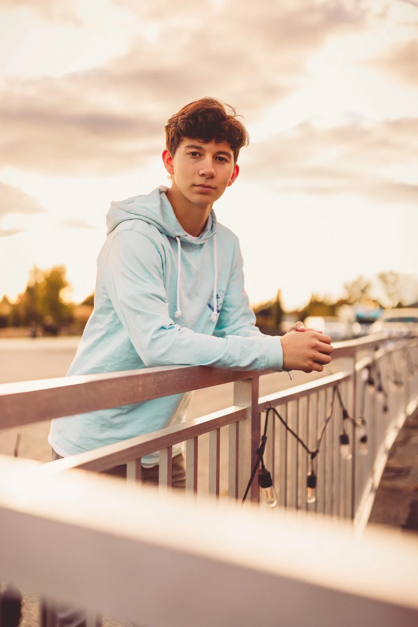A young man with curly dark hair leaning on a railing outdoors during sunset, wearing a light blue hoodie, looking at the camera.