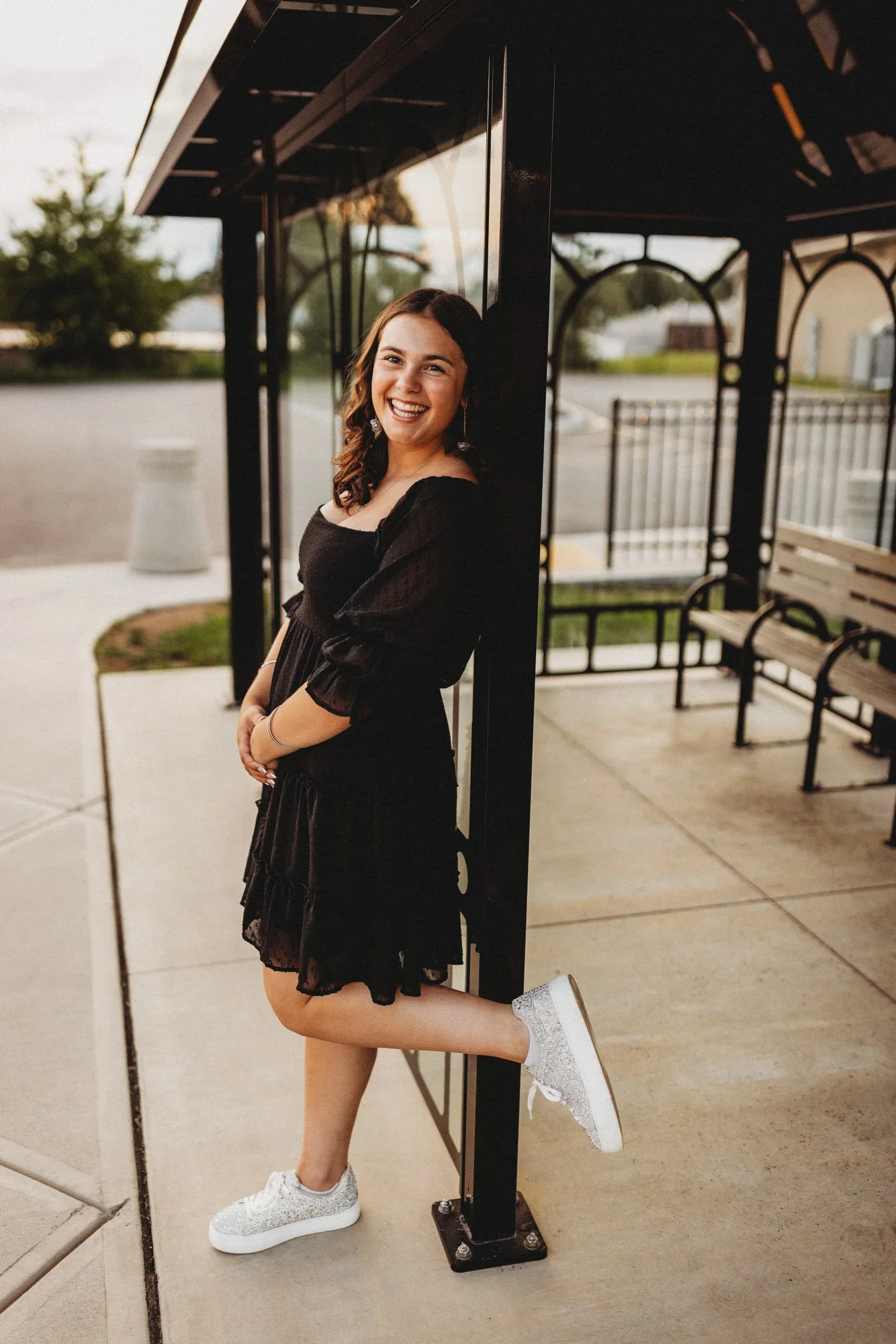 A young woman in a black dress with silver glitter sneakers, smiling and leaning against a black structure outside on a sidewalk, with benches and a railing visible behind her.