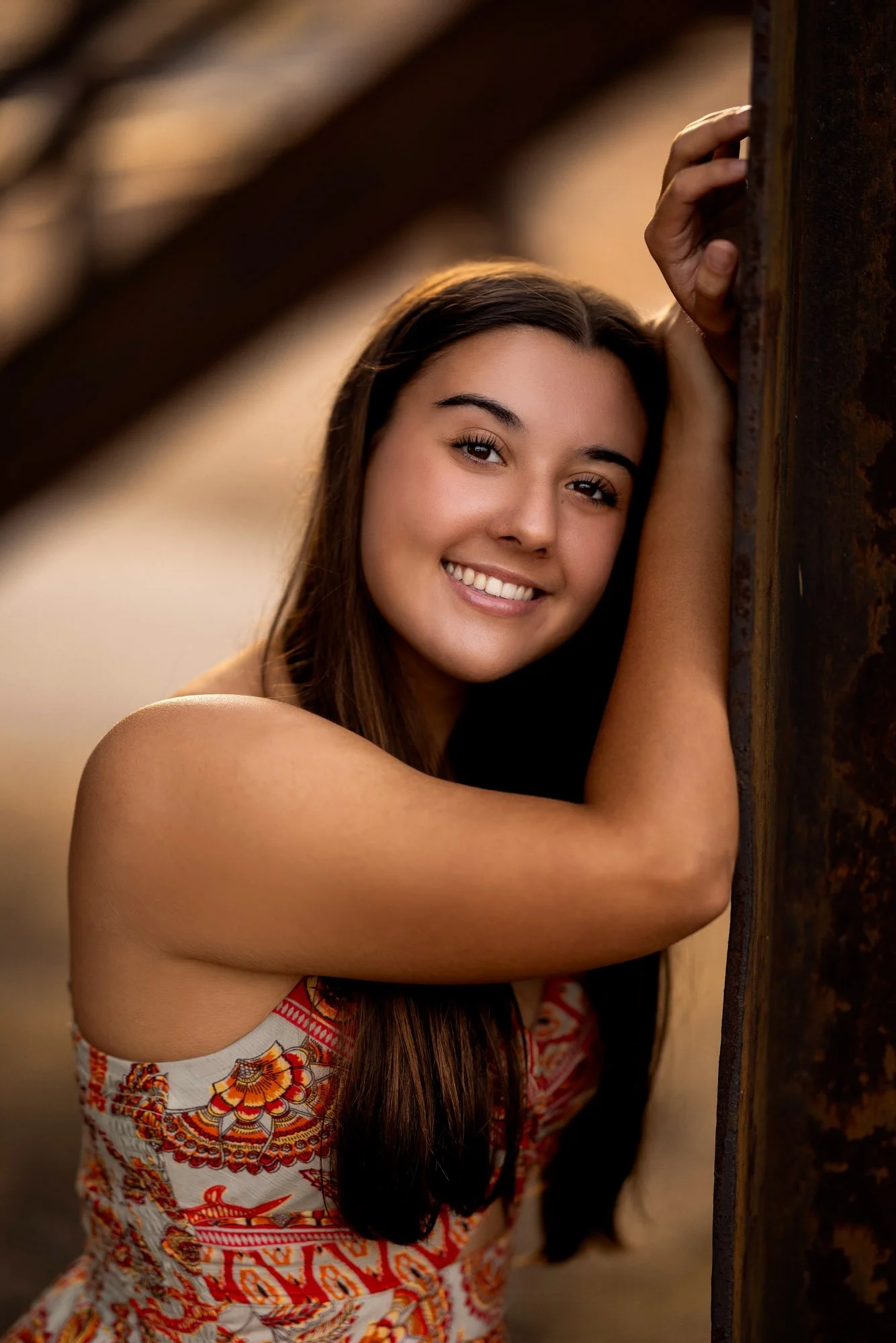 A young woman with long brown hair, smiling, leaning against a rusty metal post with her arm resting on it, outdoors during sunset.