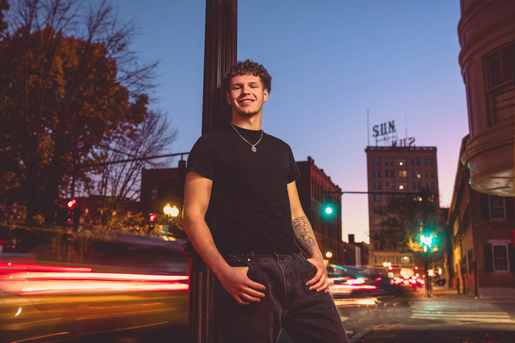 A young man with curly hair, wearing a black t-shirt and jeans, leaning against a lamppost on a city street at dusk, with streaks of car lights in the background.