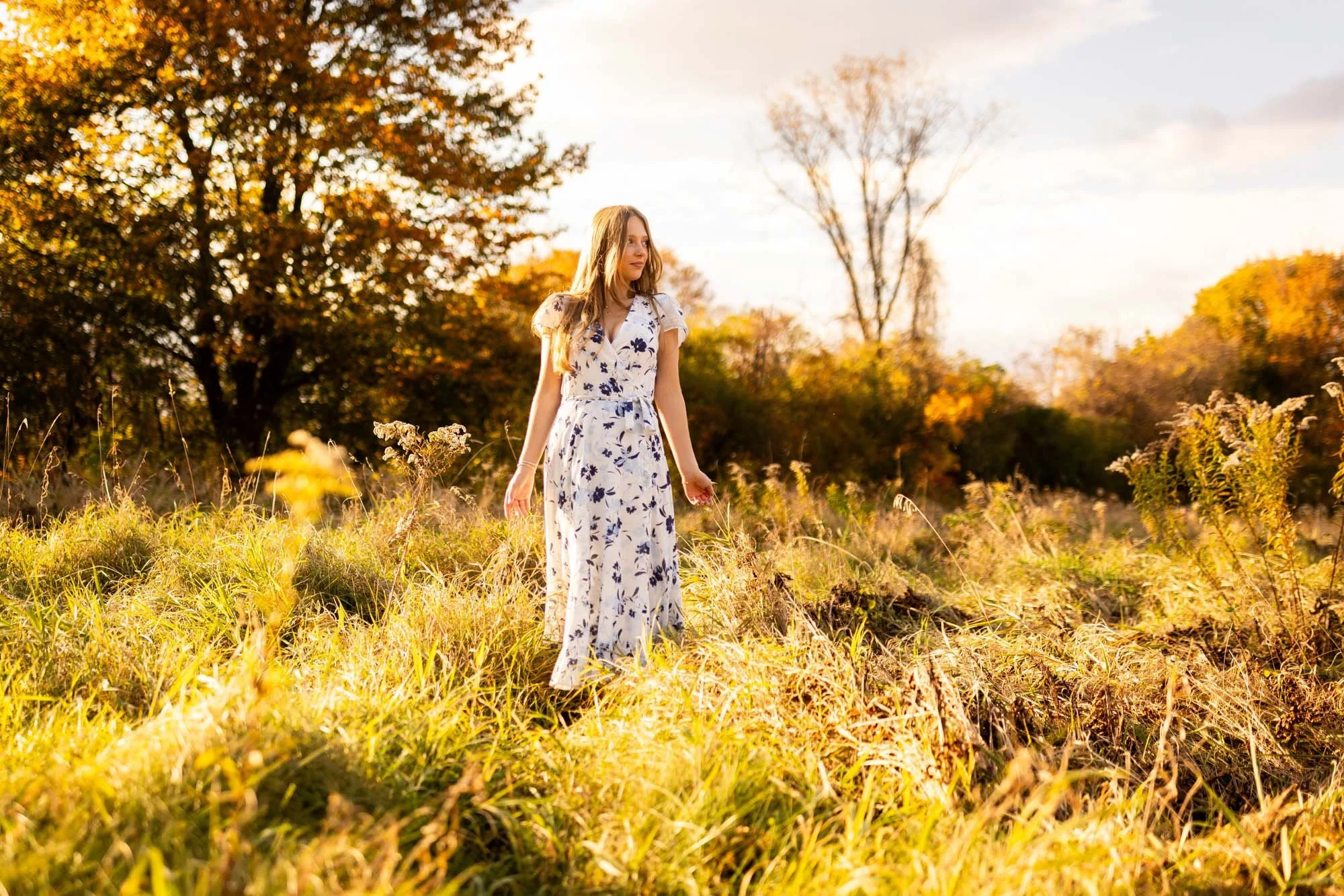 A young woman in a white floral dress walking through a grassy field during autumn, with colorful trees in the background and warm sunlight.