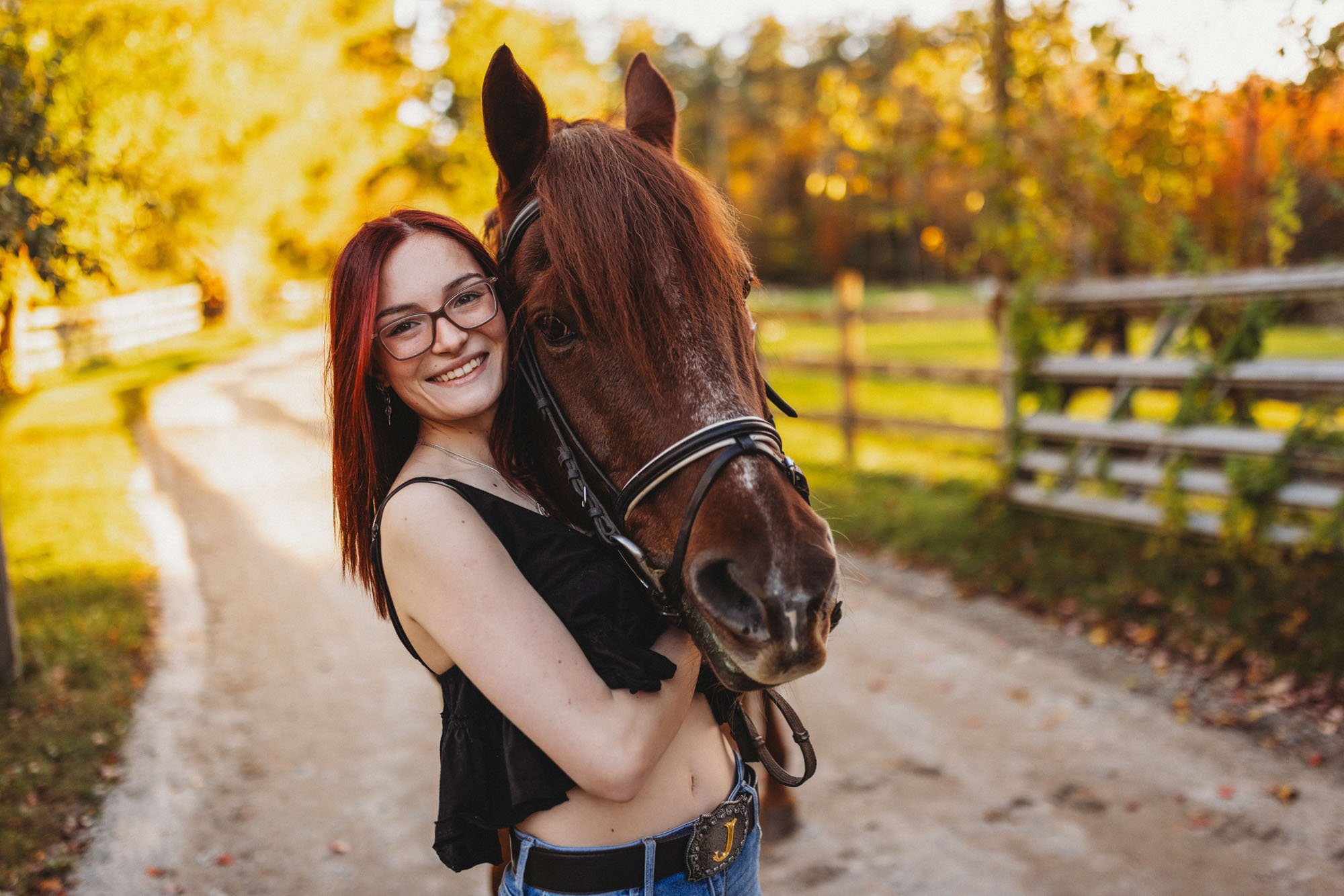 A young woman with red hair and glasses smiling while holding a brown horse with a white marking on its nose on a dirt path surrounded by autumn trees.