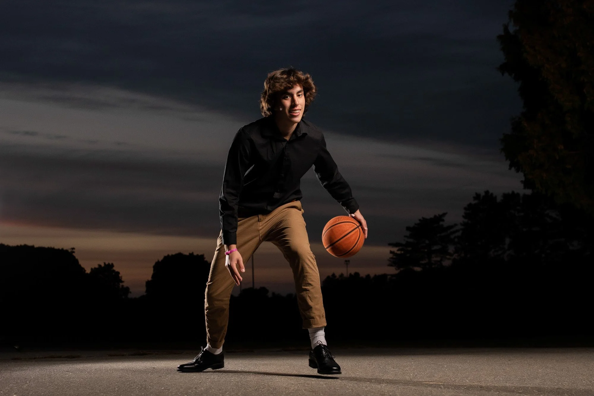 Young man with curly hair wearing a black shirt and khaki pants, holding a basketball in a dribbling position on an outdoor court at dusk, with trees and darkening sky in the background.