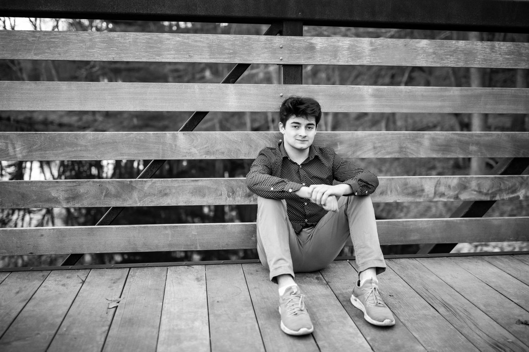 Black and white photo of a young man sitting on a wooden bench outdoors, wearing a patterned shirt, light pants, and sneakers, with a relaxed expression.