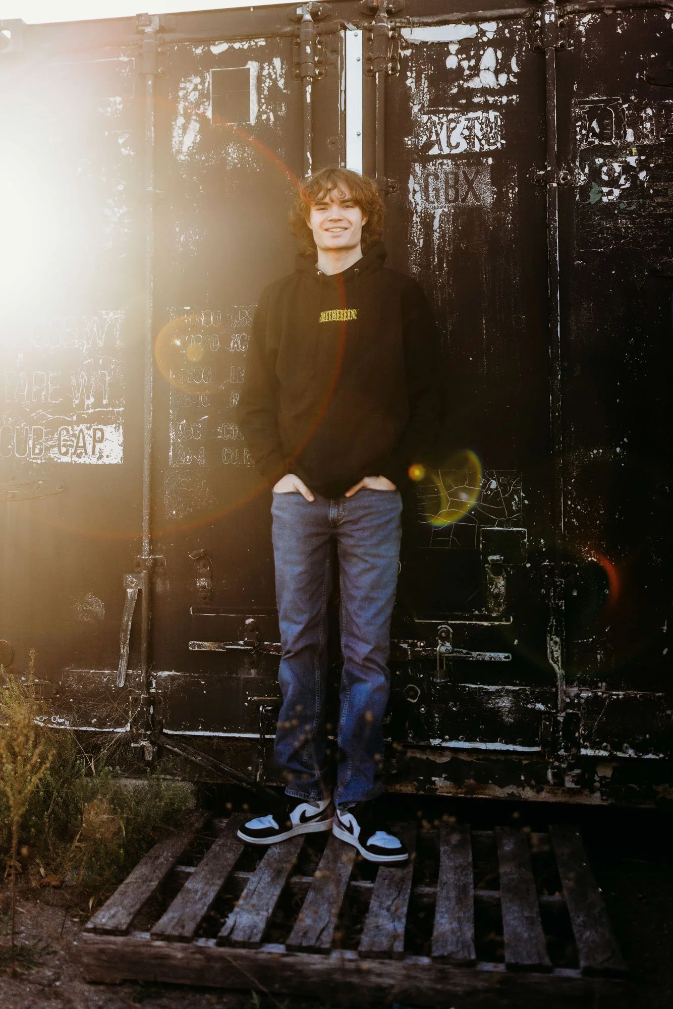 A young man with curly hair wearing a black hoodie, jeans, and Nike sneakers, standing in front of a weathered black metal door with graffiti, illuminated by sunlight creating lens flare effects.