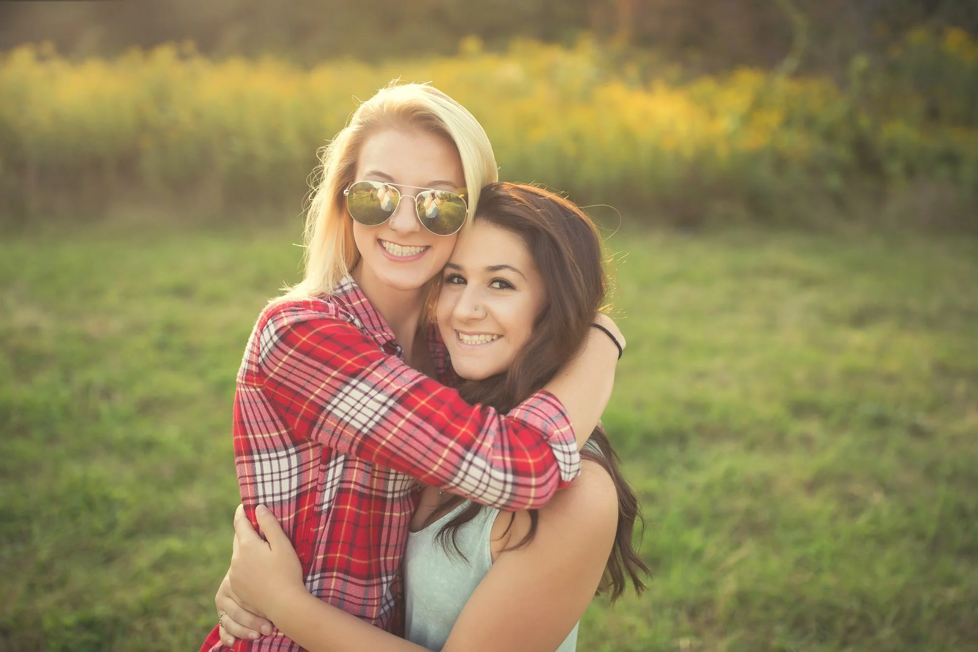 Two smiling young women hugging outdoors in a grassy field during sunset, one wearing sunglasses and a red plaid shirt, the other with long dark hair and a light blue top.