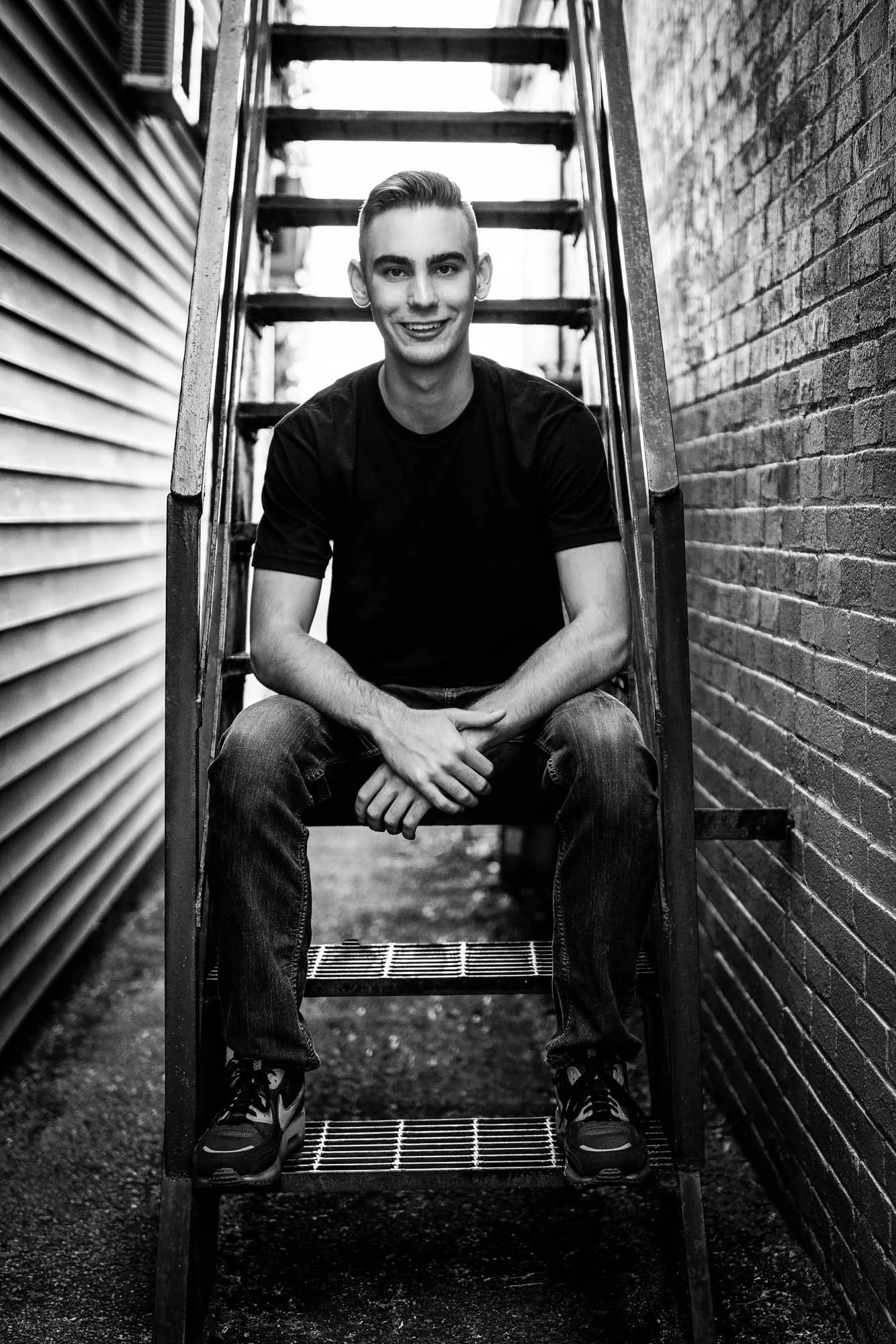 A young man with short hair, wearing a black t-shirt and jeans, sitting on metal stairs between two brick walls, smiling at the camera in black and white.