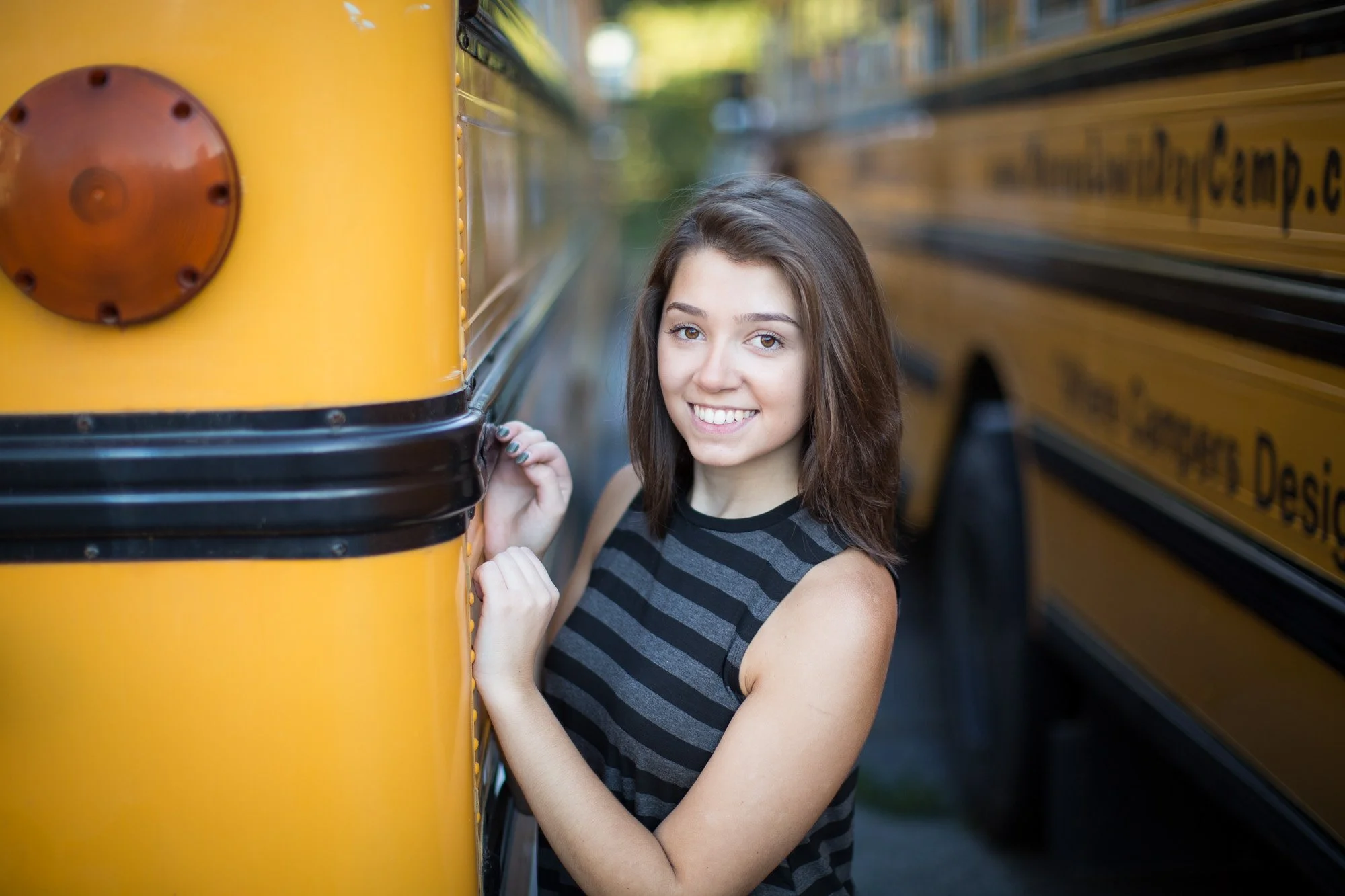 Young woman with shoulder-length brown hair smiling while standing between two yellow school buses.