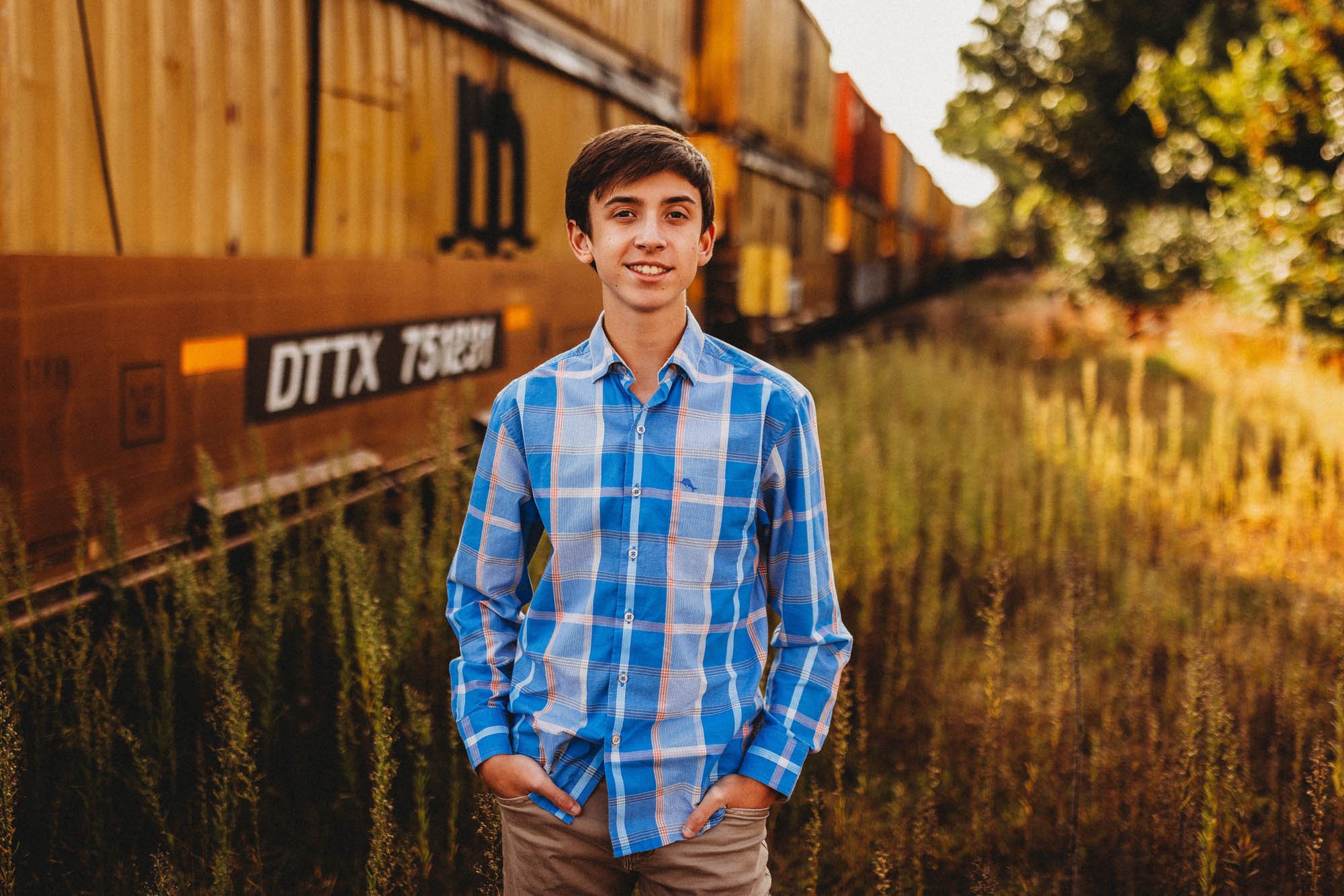A young man in a blue plaid shirt standing outdoors near a train with colorful freight cars, autumn trees, and a reflection in the water.