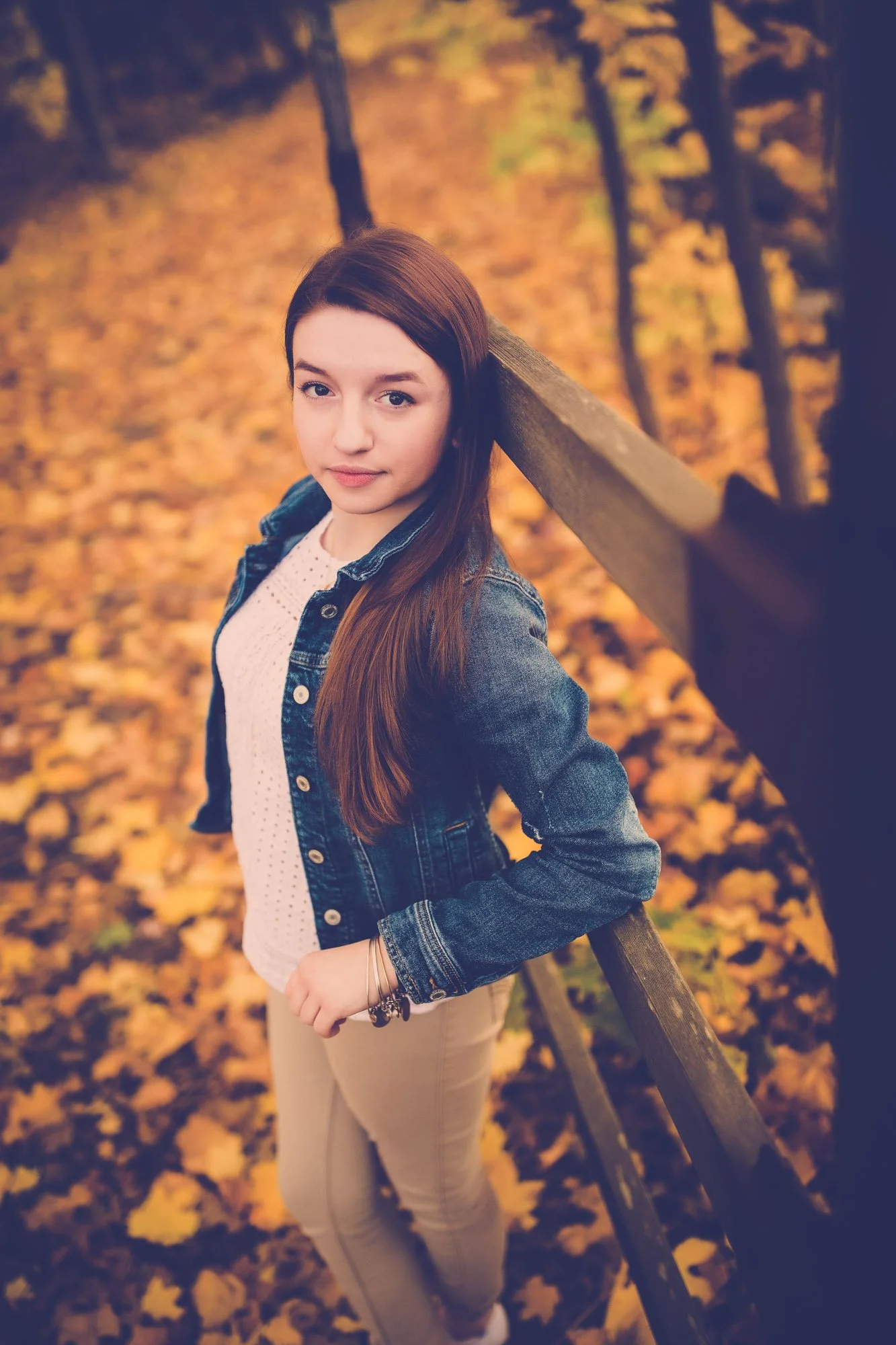 A young woman with long brown hair standing outdoors on an autumn day, leaning against a wooden fence, with fallen yellow leaves covering the ground behind her.