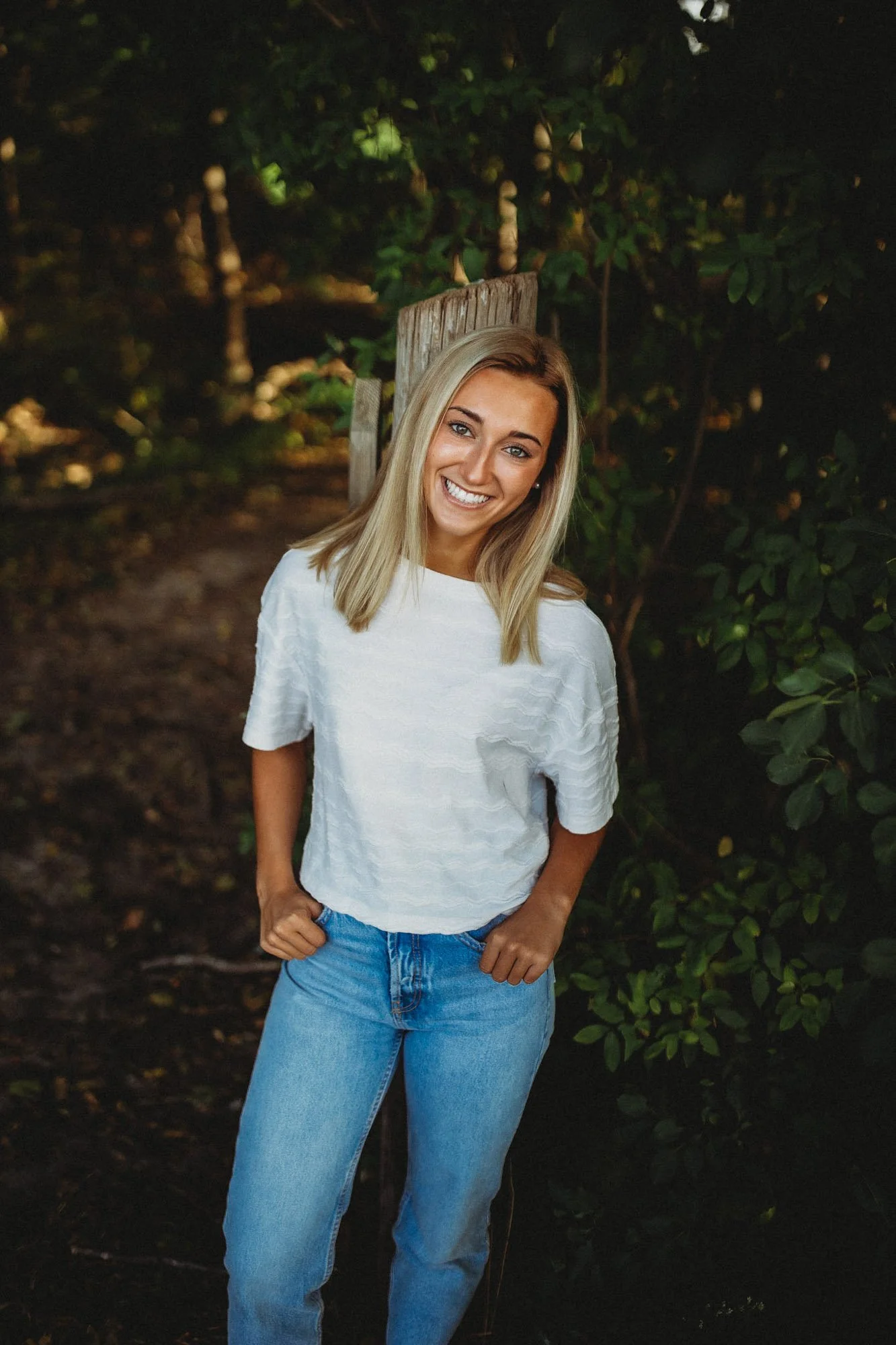A young woman with blonde hair smiling outdoors, standing near a wooden post surrounded by green foliage, wearing a white textured T-shirt and light blue jeans.