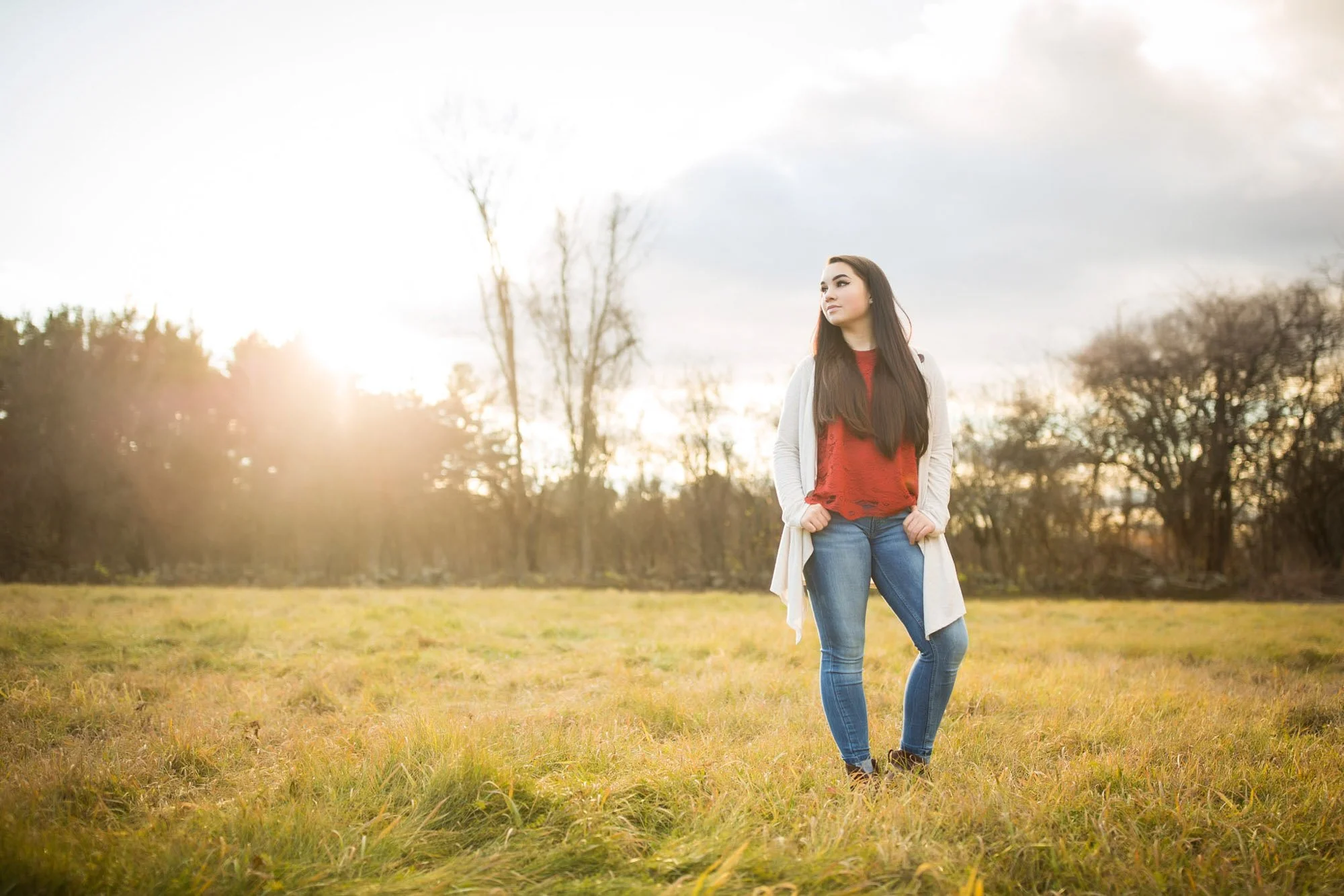 Young woman standing in a grassy field with trees in the background and sunlight shining through on a cloudy day.