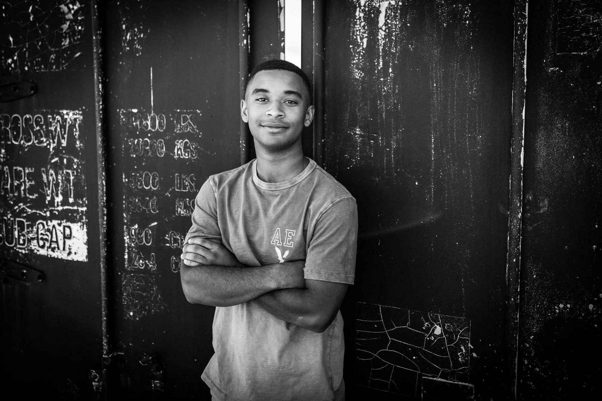 A young man standing with arms crossed in front of a black painted wall with graffiti and weathered paint, looking at the camera, in an urban setting.