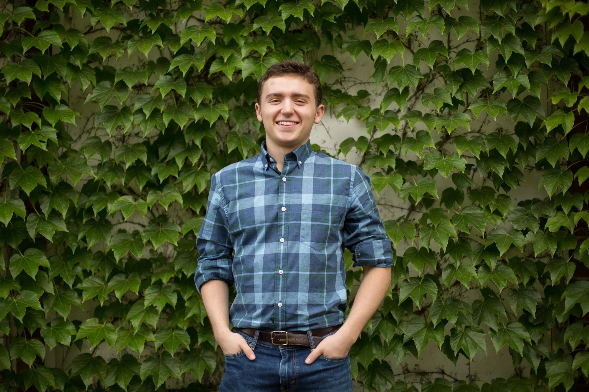 A young man with short brown hair wearing a blue plaid shirt and jeans standing outdoors in front of a wall covered in green ivy.