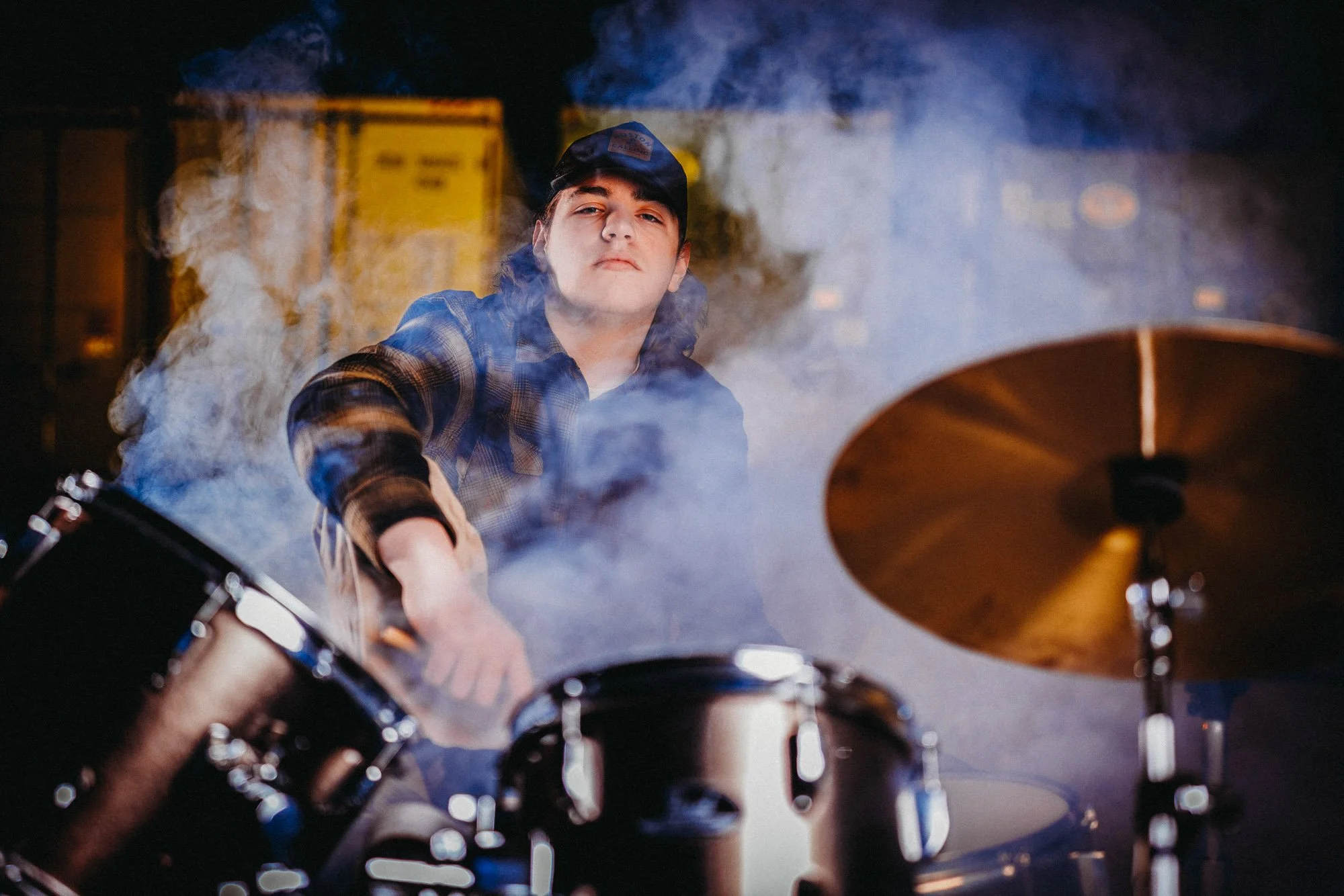 A young man wearing a black cap and a plaid shirt is playing drums, surrounded by smoke with a focused expression.