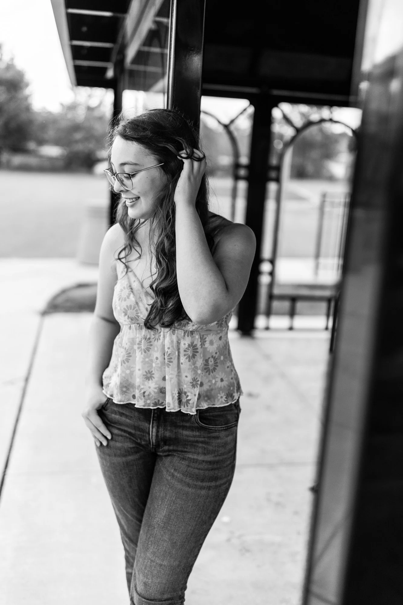 A young woman with long wavy hair and glasses, smiling while standing outside near a building, in black and white.