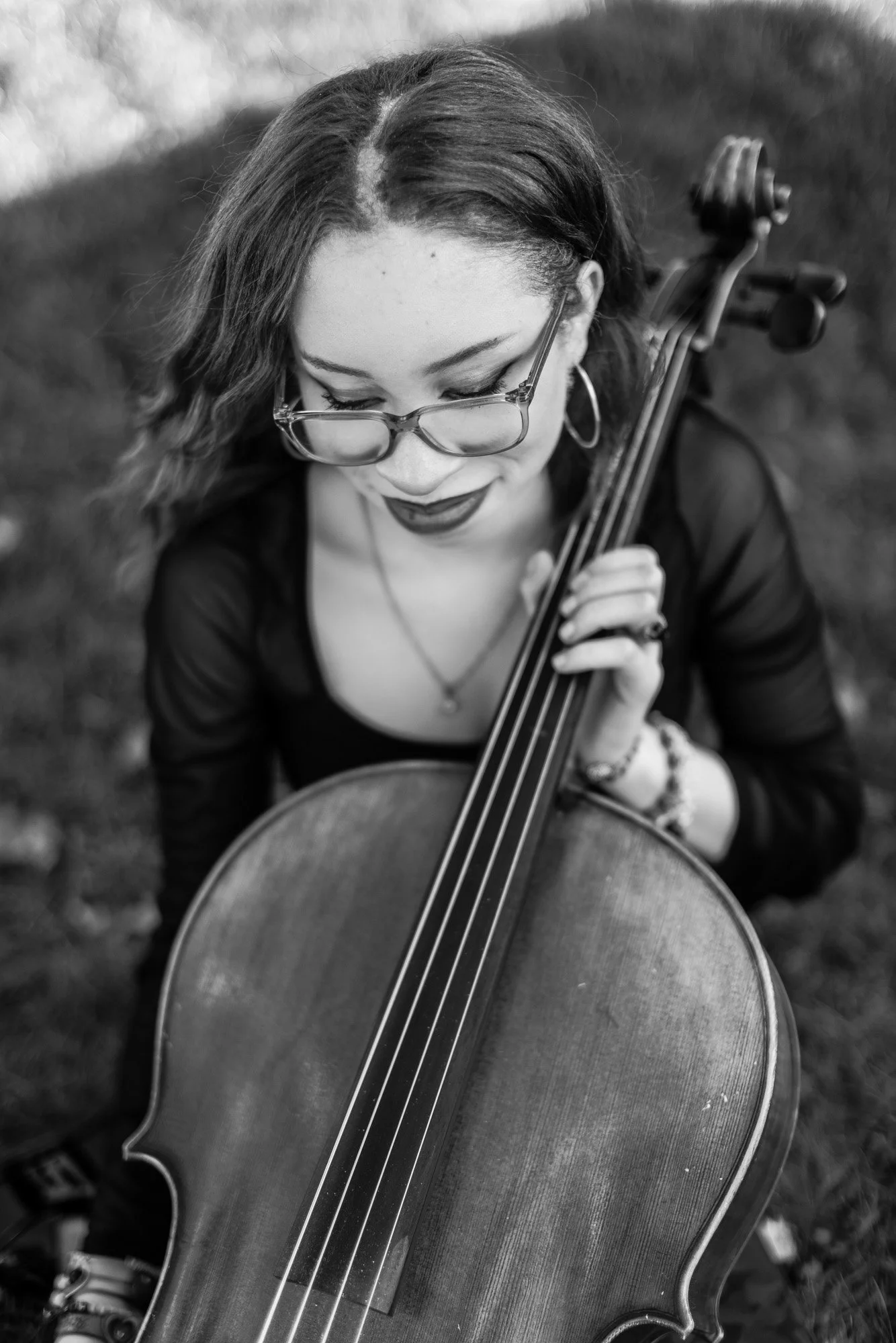 A black-and-white photo of a woman with glasses, dark lipstick, and hoop earrings, sitting outdoors with her cello resting against her body, looking down at it.