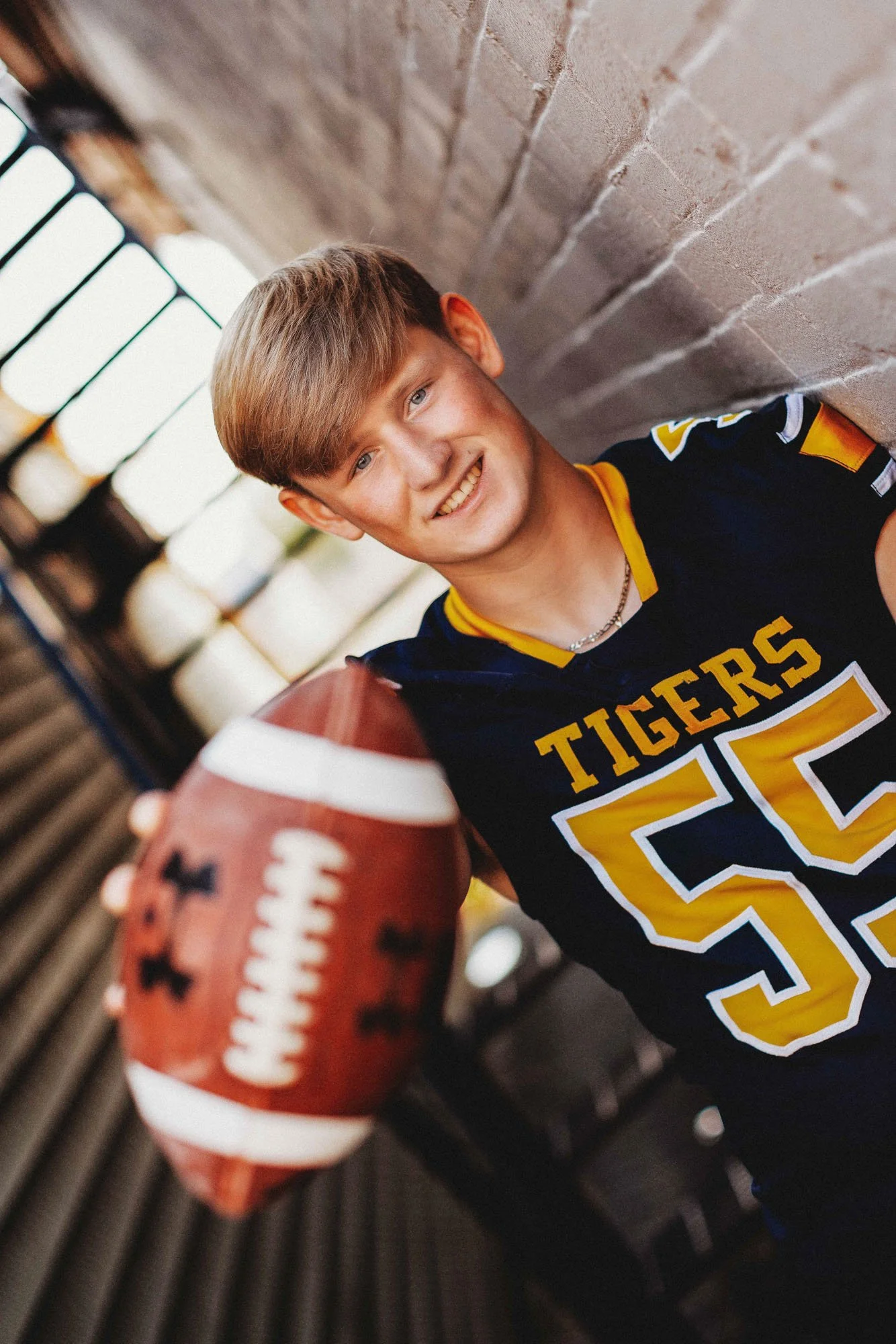Young man in a black and yellow football jersey holding a football, smiling, leaning against a brick wall.