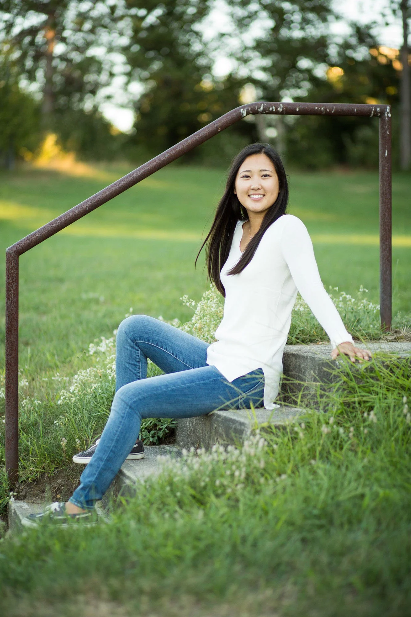 Young woman sitting on a concrete step outdoors, smiling, with grass and trees in the background.